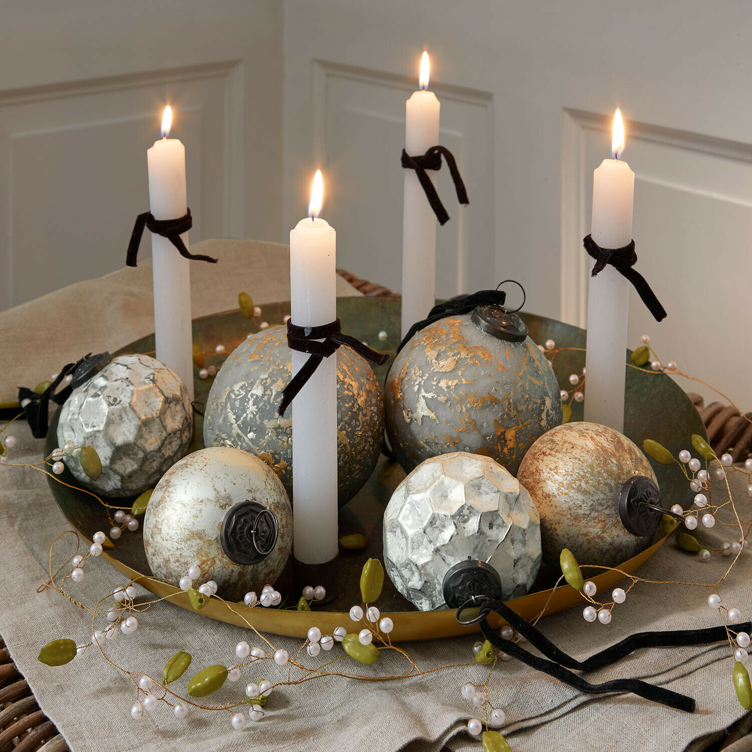 Goldene Metallplatte mit weißen Kerzen und dekorativen Kugeln in Grau- und Beigetönen auf Leinentuch