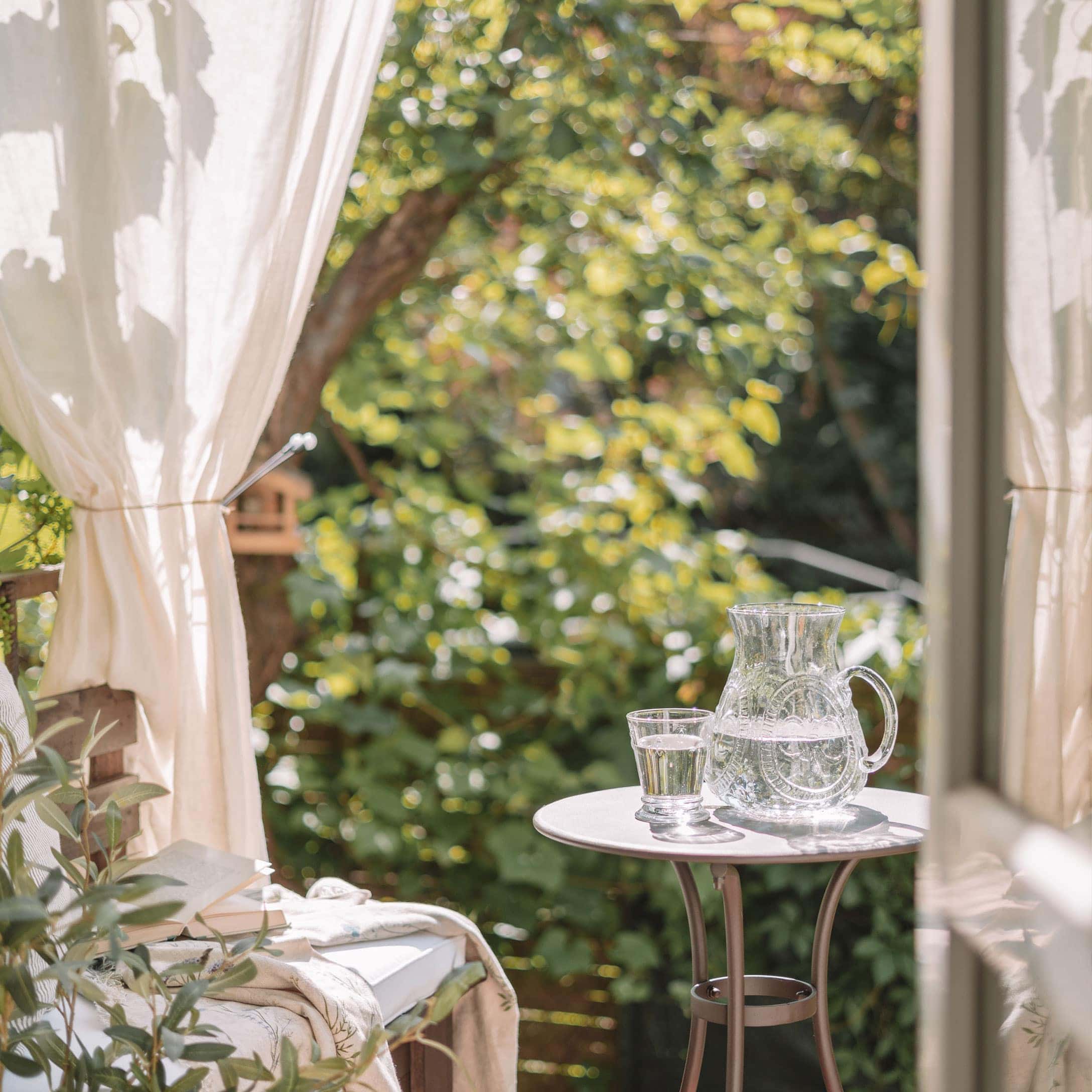 Glaskrug und Glas mit Wasser auf weißem Tisch auf Terrasse mit grünem Pflanzenhintergrund