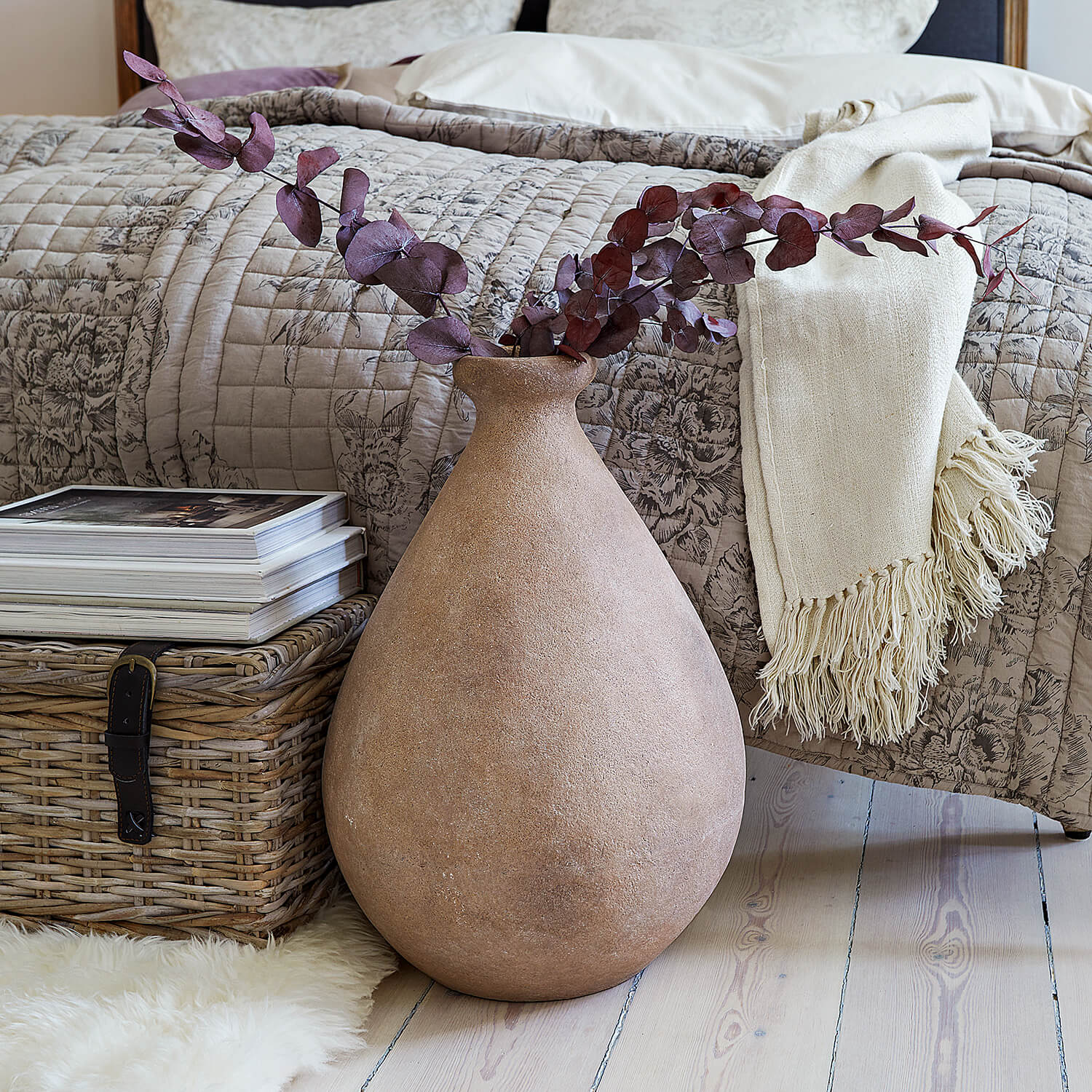 Große beige Keramikvase mit getrockneten Zweigen vor Bett und Korb aus Weide