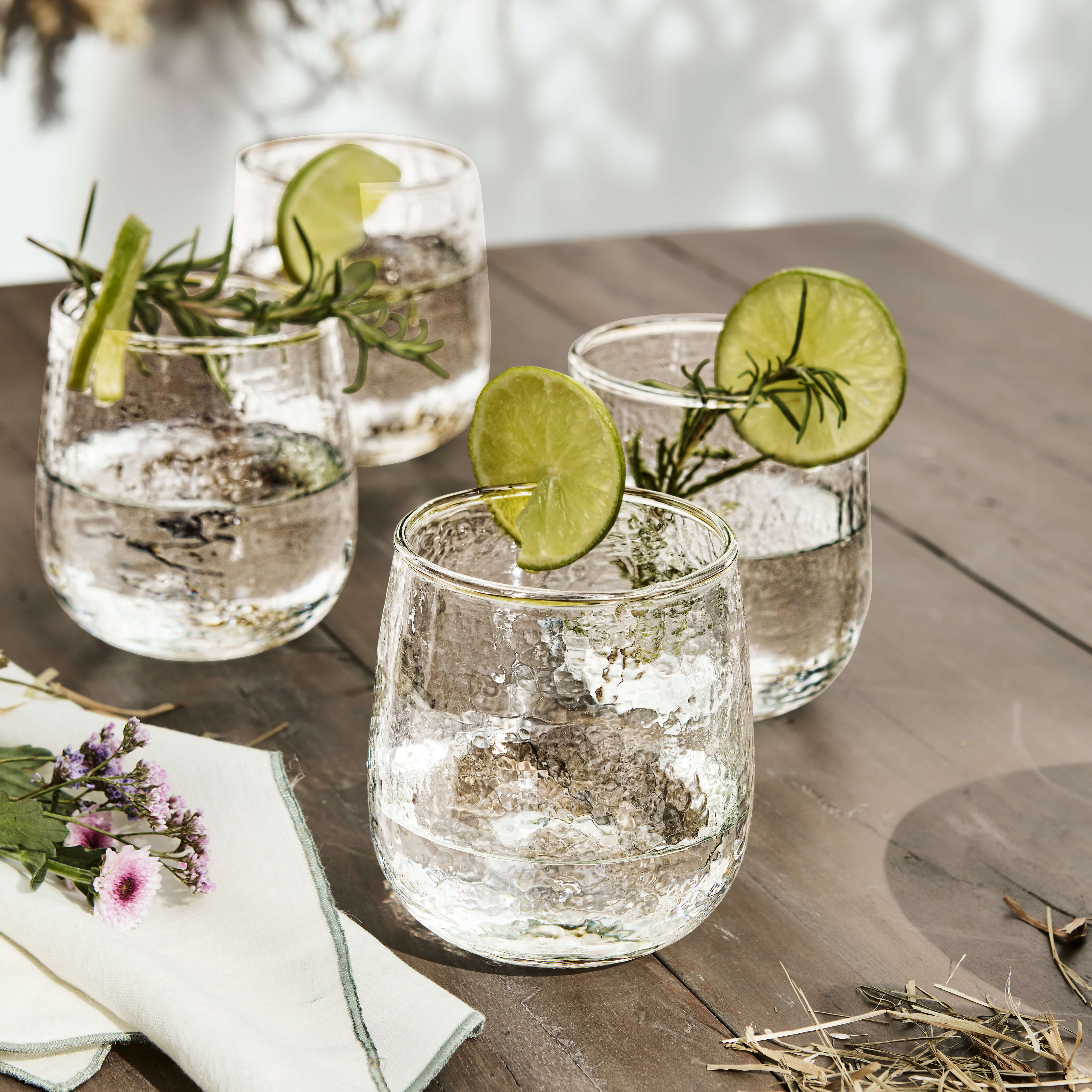 Vier strukturierte Gläser mit Wasser, Limettenscheiben und Rosmarinzweigen auf Holztisch arrangiert