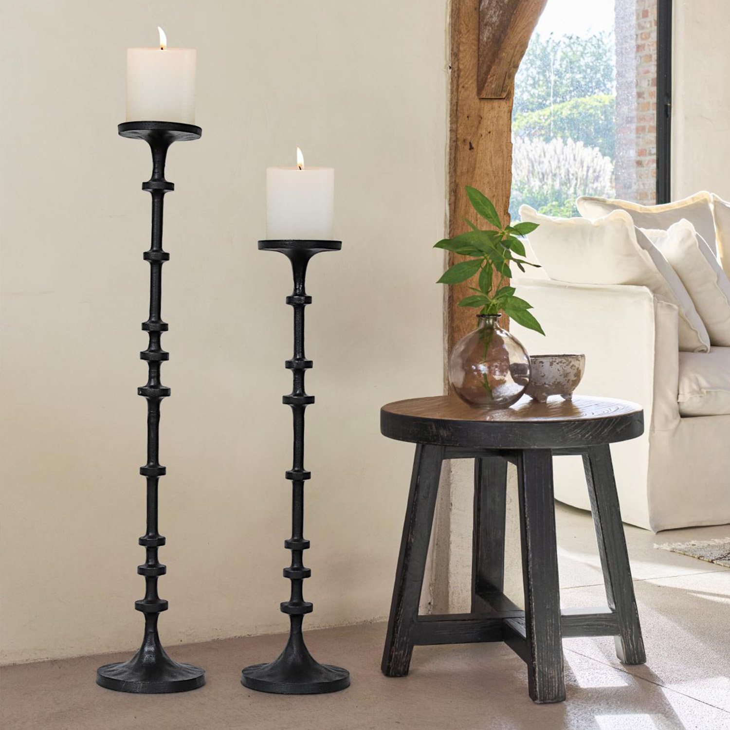 Zwei schwarze Metall-Kerzenhalter mit weißen Kerzen neben einem Holzhocker und Sofa im Wohnzimmer