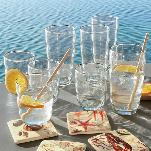 Gläser aus klarem Glas mit Zitronenscheiben und Holzrührstäbchen, maritime Untersetzer am Wasser
