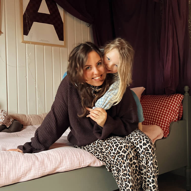 Frau im braunen Pullover und Leopardenhose sitzt mit Kind im hell karierten Hemd auf Bett