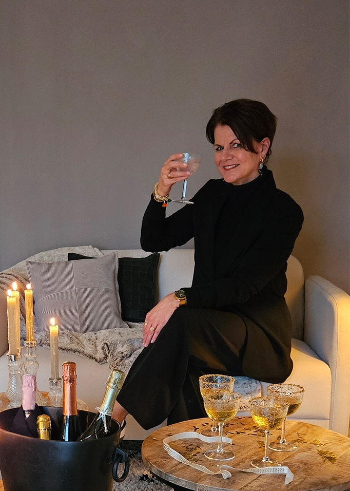 Frau in schwarzer Kleidung sitzt auf hellem Sofa mit Glas, Kerzen und Champagnerflaschen daneben