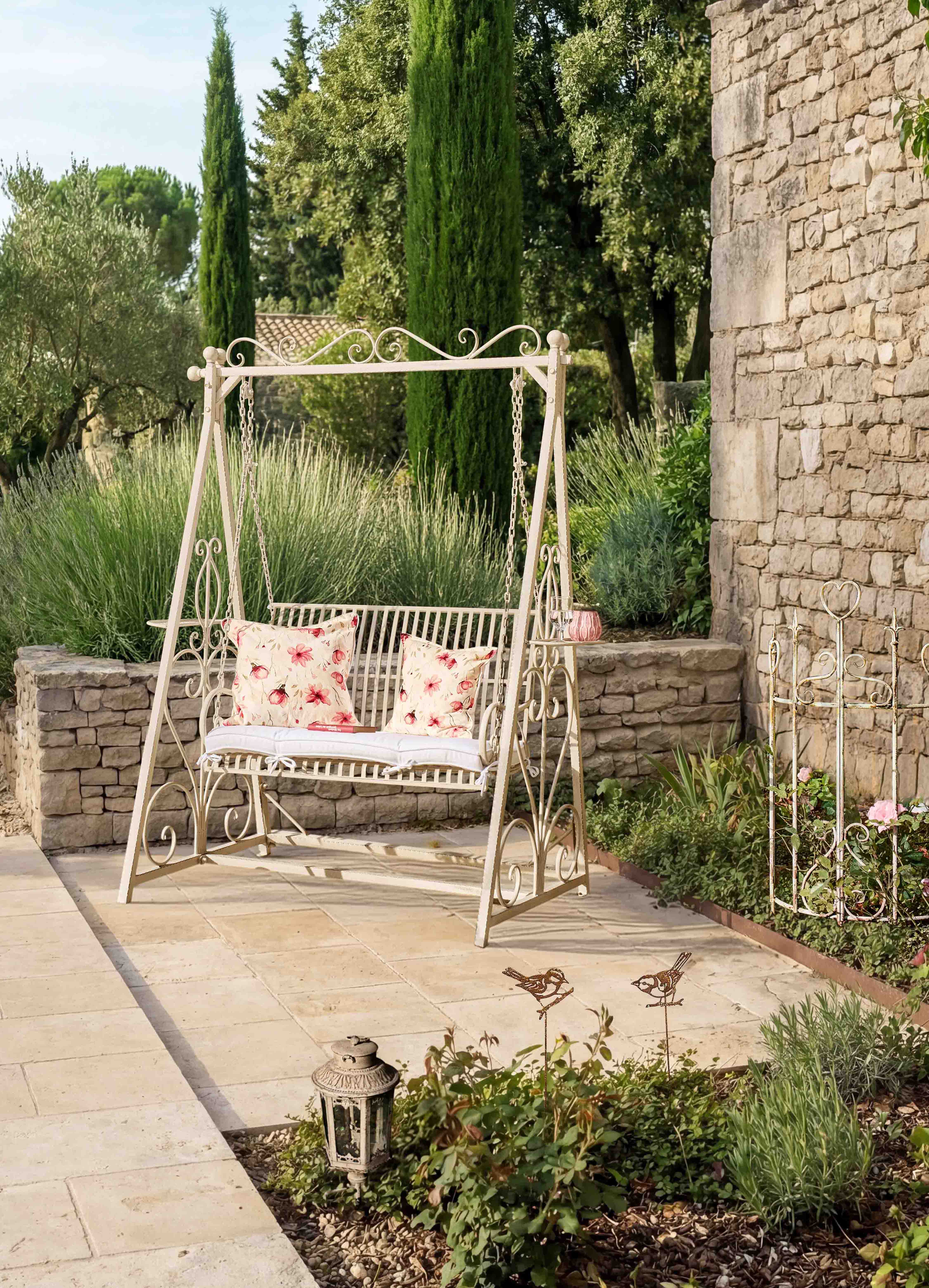 Metall-Gartenschaukel in Creme mit floral gemusterten Kissen auf Terrasse mit Natursteinmauer