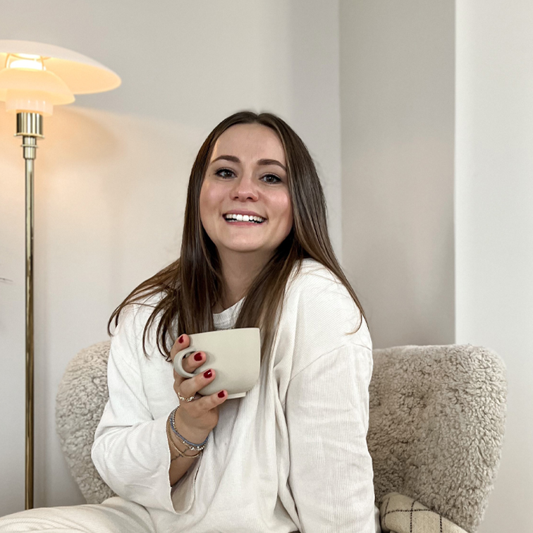 Lächende Frau im weißen Pullover sitzt auf weichem Sessel und hält eine beige Tasse