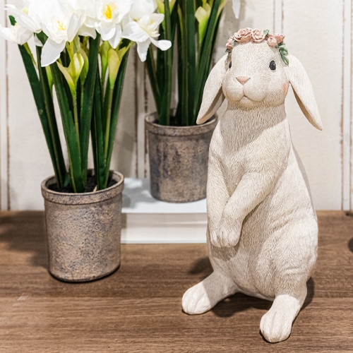 Dekorativer weißer Hase mit Blumenkranz auf Holztisch neben Töpfen mit weißen Blumen
