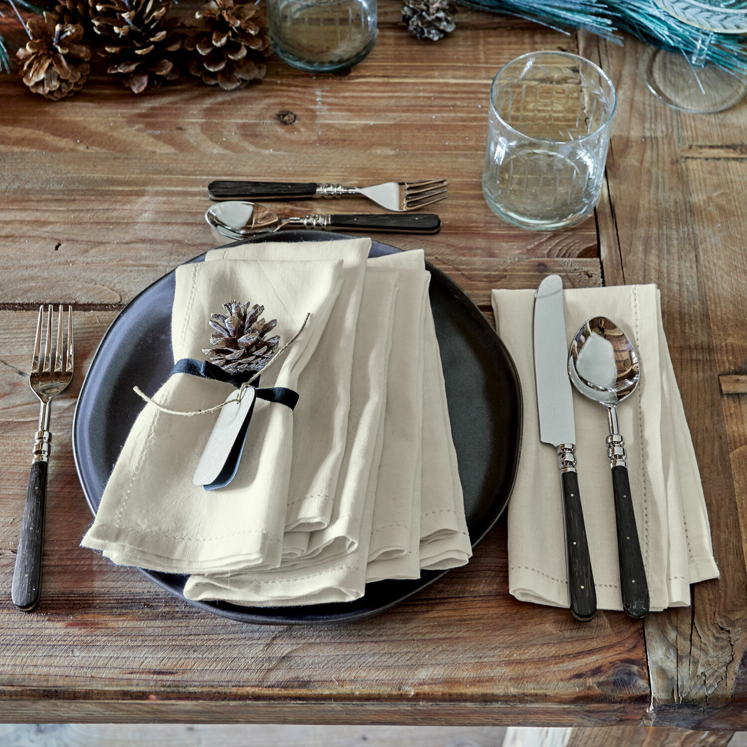 Rustikaler Holztisch mit schwarzem Teller, cremefarbenen Leinenservietten und Besteck mit schwarzen Griffen