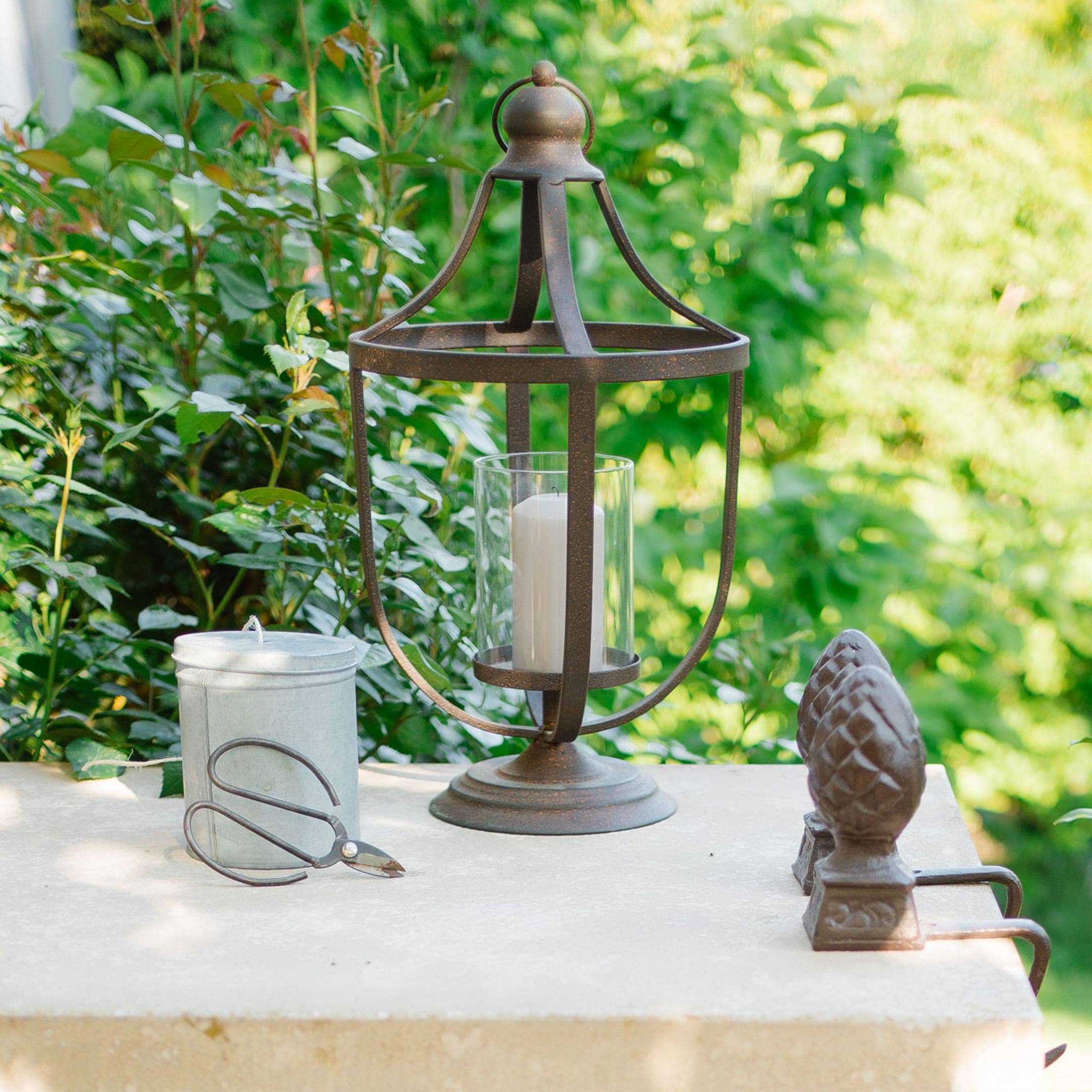 Metalllaterne mit weißer Kerze und Gartenschere auf hellem Tisch vor grünem Hintergrund