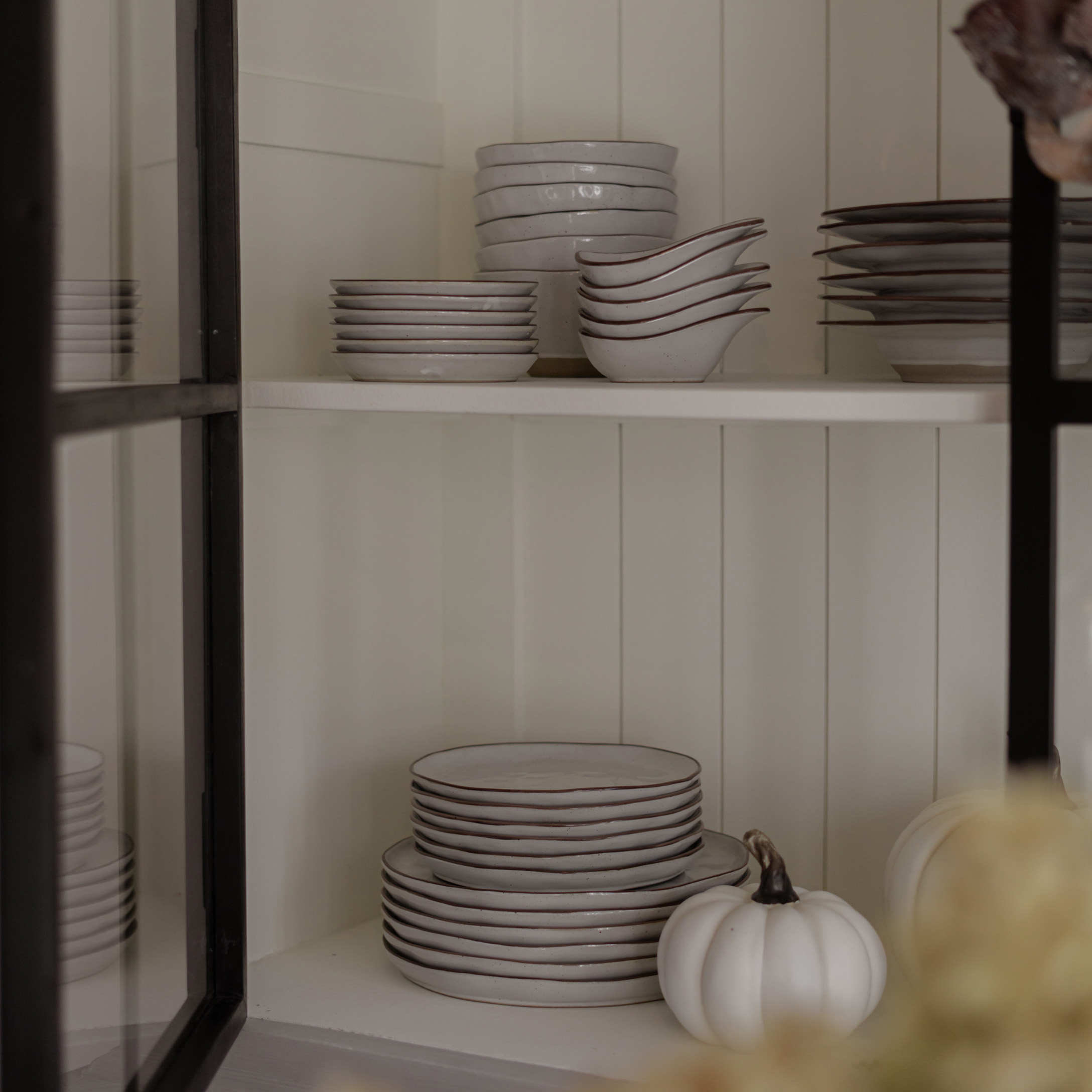 Weißes Keramikgeschirr mit braunem Rand auf Regal, dekorativer weißer Kürbis daneben, rustikaler Stil