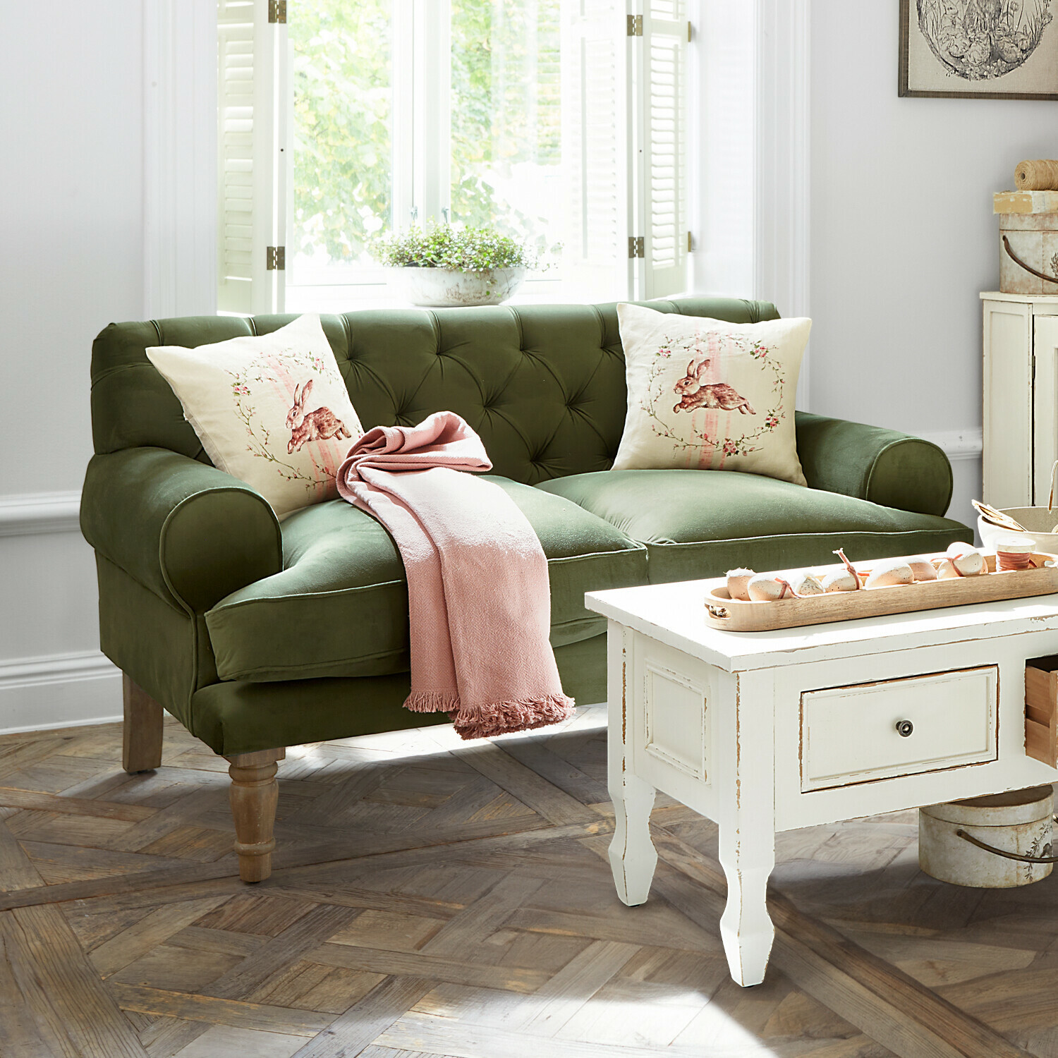 Sofa Carme Grünes Samtsofa mit Hasenmotiv-Kissen und rosa Decke in hellem Wohnzimmer