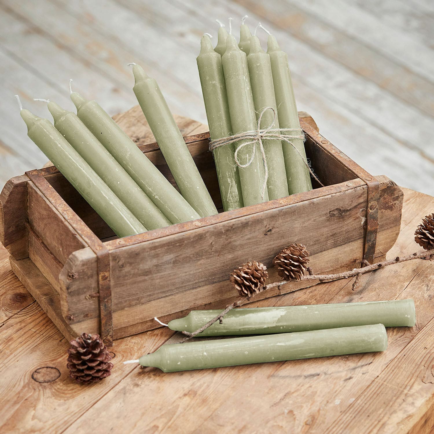 Kerze 12er Set Bauceau Set aus grünen Wachskerzen in einer Holzkiste auf rustikalem Holztisch mit Kiefernzapfen