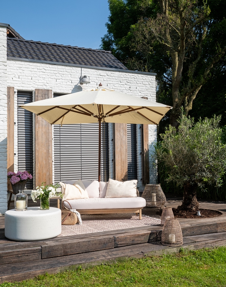 Terrasse mit weißer Ziegelwand, beiges Sofa mit Kissen und cremefarbenem Sonnenschirm auf Holzdeck