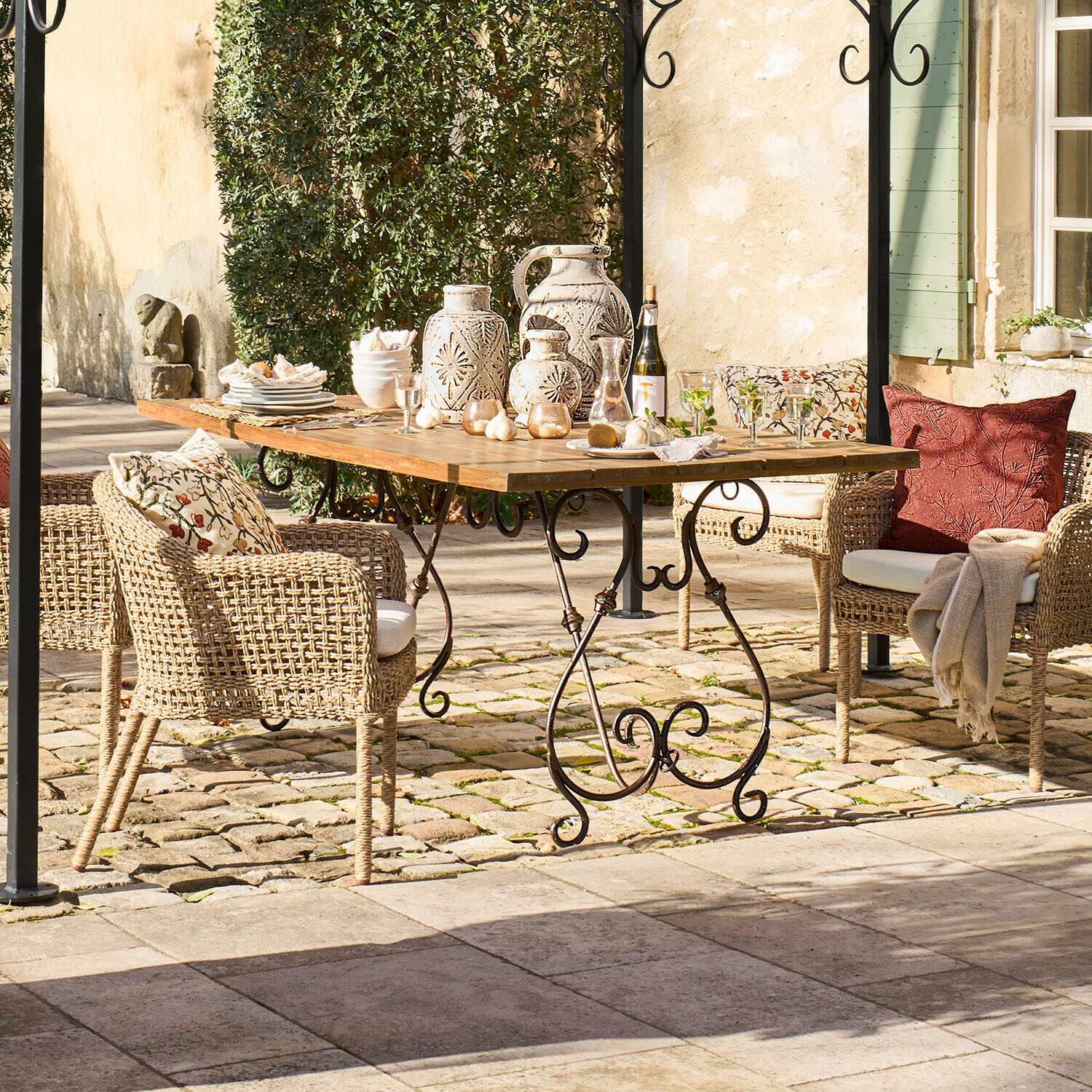 Tisch Delbridge Gartentisch mit Holzplatte und Metallbeinen, umgeben von Rattanstühlen mit Kissen und Keramikdekorationen