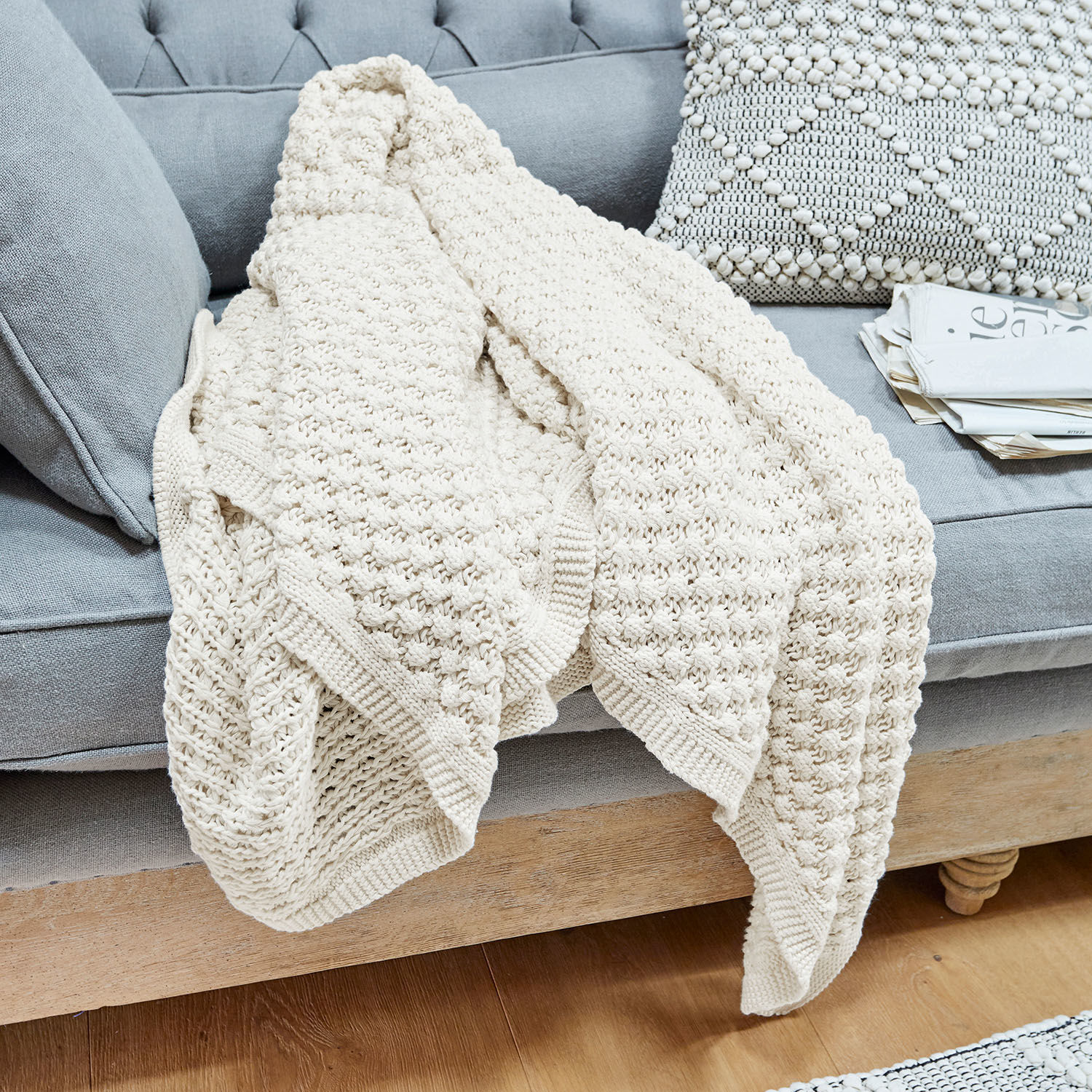 Plaid Tremes Beige gestrickte Baumwolldecke auf grauem Sofa mit dekorativem Kissen und Zeitungen