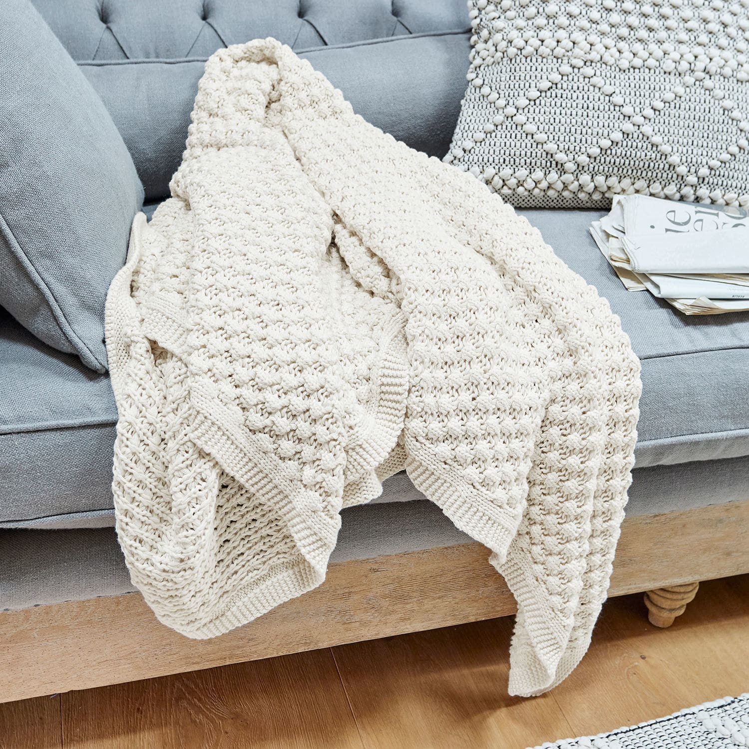 Plaid Tremes Beige gestrickte Baumwolldecke auf grauem Sofa mit dekorativem Kissen und Zeitungen