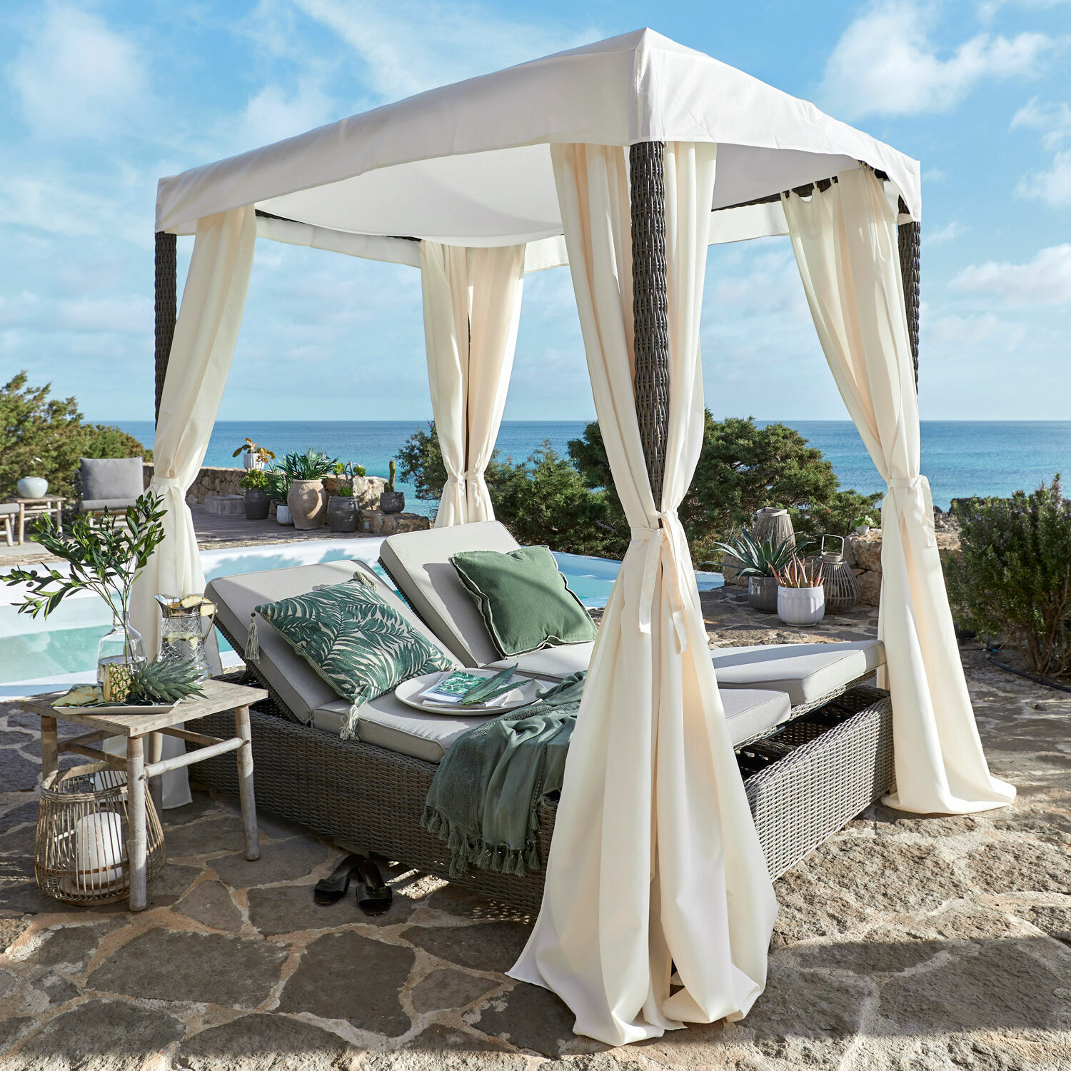 Gartenlounge Hereford Gartenliege aus Rattan mit cremefarbenem Baldachin auf Steinboden mit Meerblick