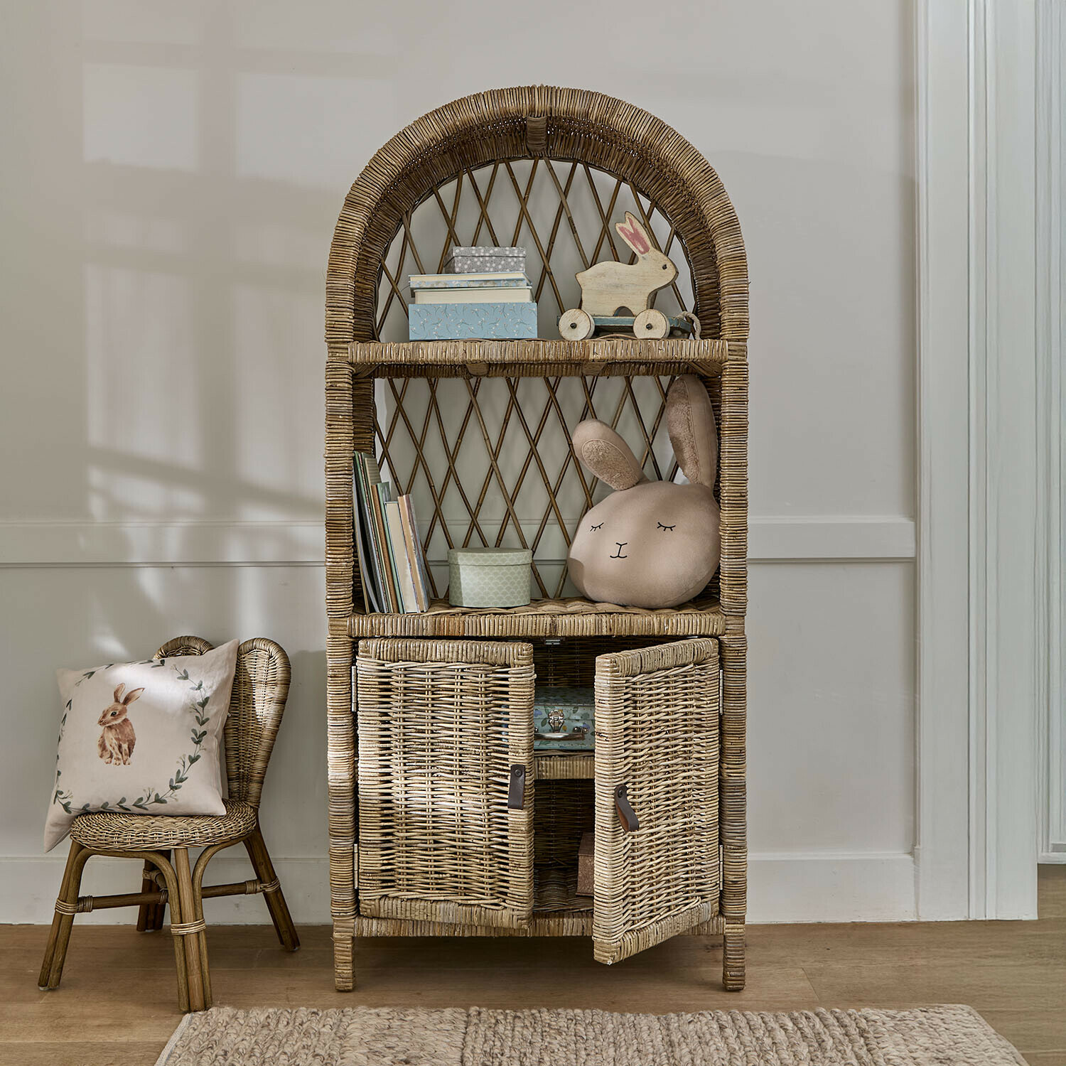 Rattanschrank mit abgerundeten Regalen und Türen in Naturfarbe, Kinderdeko und Hasenkissen