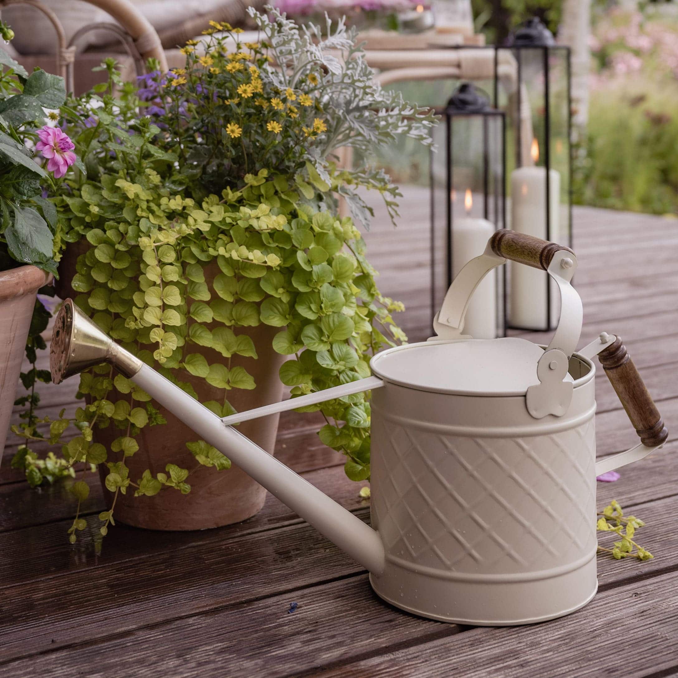 Beige Metall-Gießkanne mit Holzgriff auf Holzterrasse neben Pflanzen und Kerzen