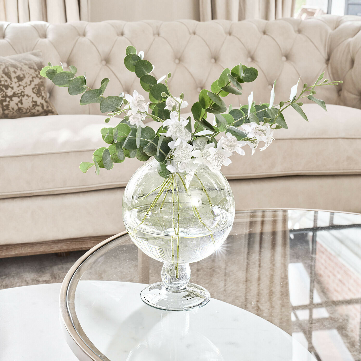 Vase Marsetta Transparente Glasvase mit grünen Blättern und weißen Blumen auf Glastisch im hellen Wohnzimmer