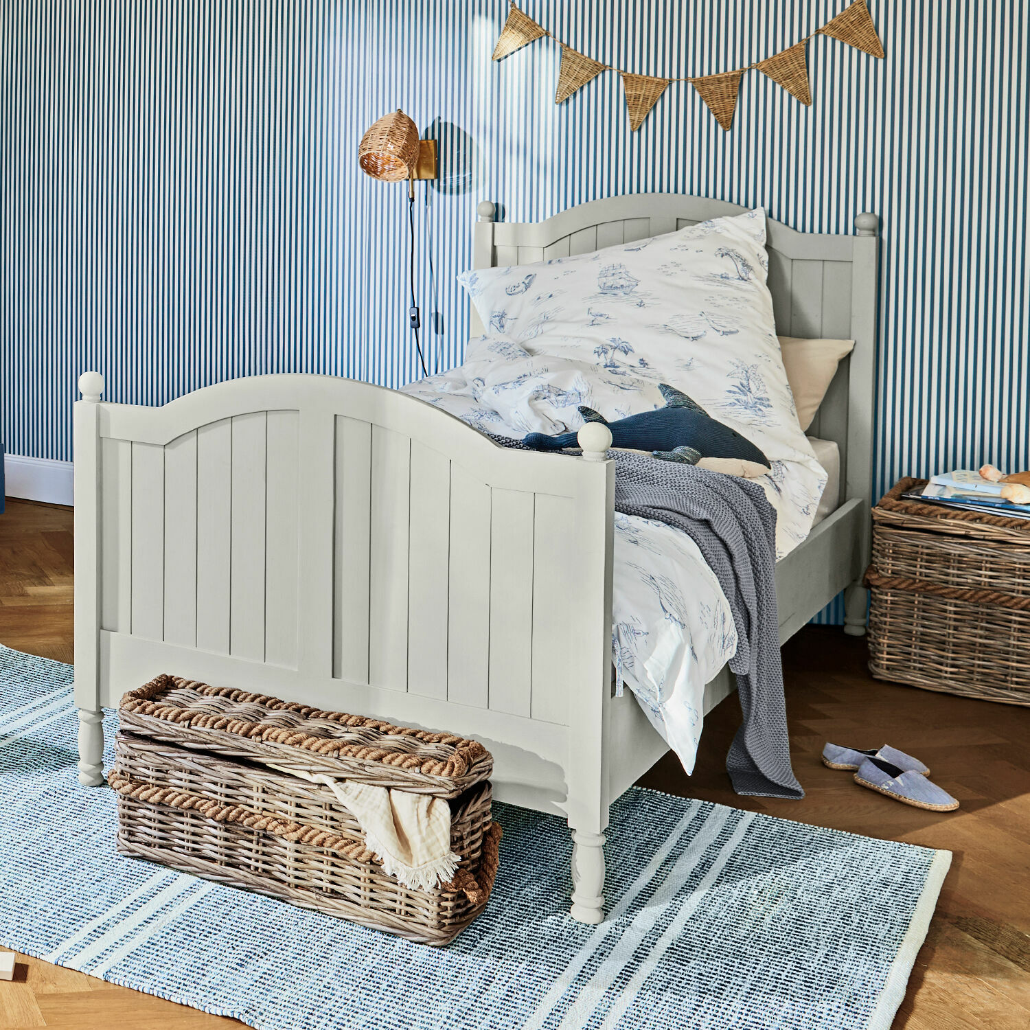 Bett Alarion Graues Kinderbett aus Holz mit maritim gemusterter Bettwäsche und Korb aus Weide auf Teppich
