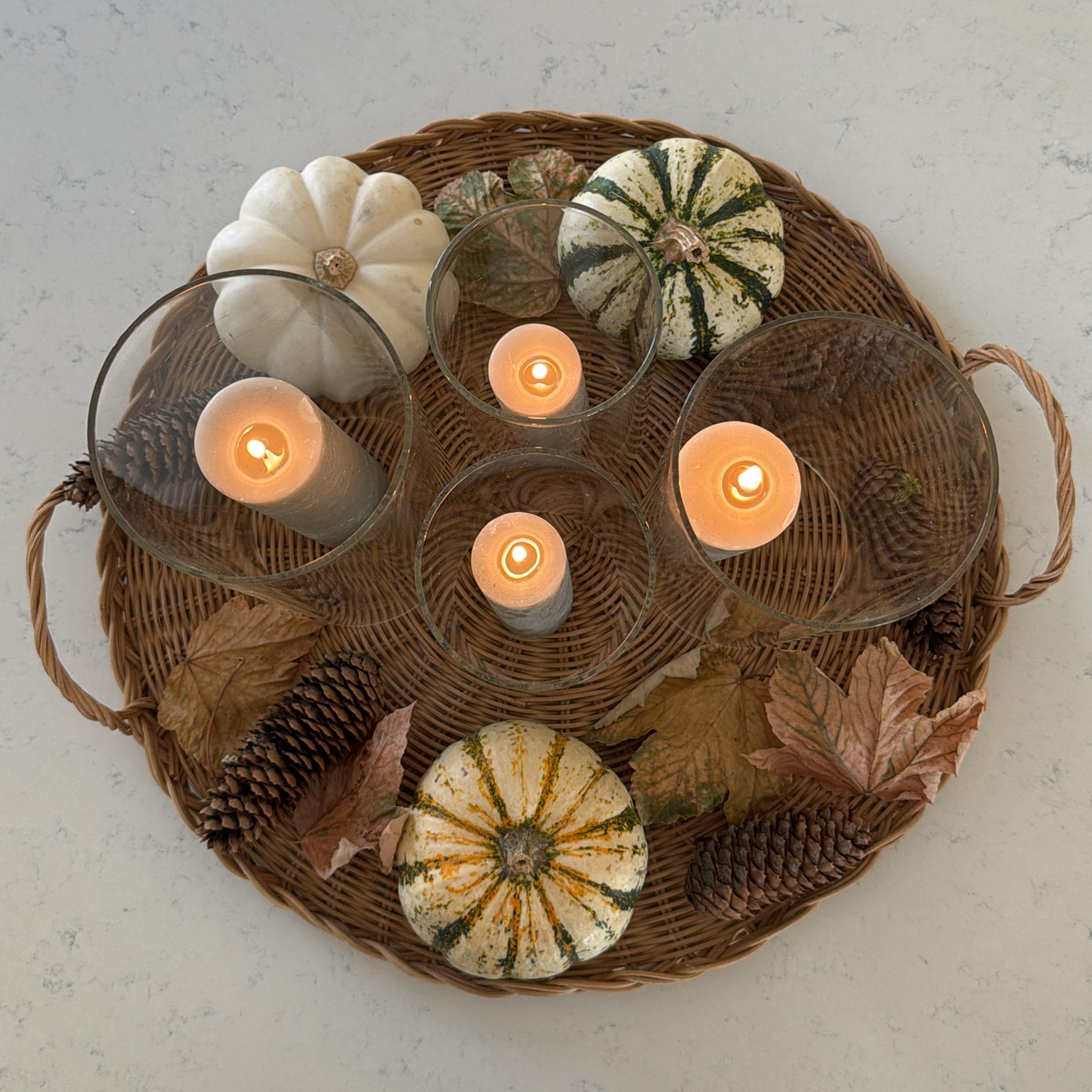 Herbstliches Ensemble mit weißen Kürbissen, Tannenzapfen und brennenden Kerzen in Glas auf einem geflochtenen Tablett