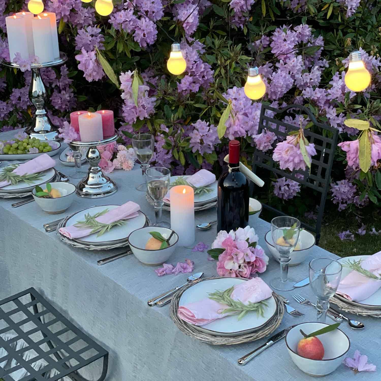 Eleganter Gartentisch mit rosa Servietten, Kerzen und Weinflasche vor lila Blüten dekoriert