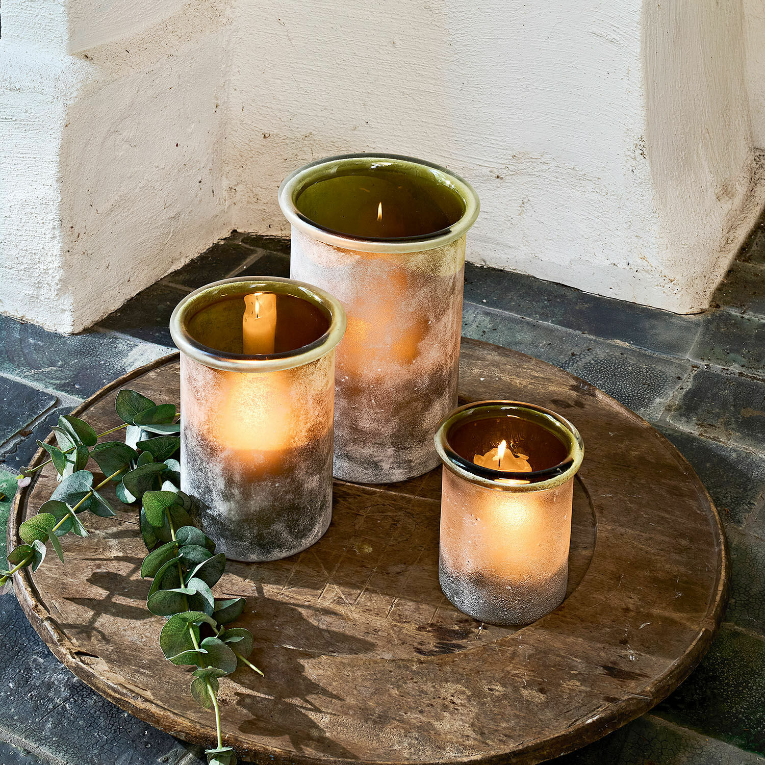 Drei matte Glaswindlichter in Braun- und Grüntönen auf rustikalem Holztablett arrangiert