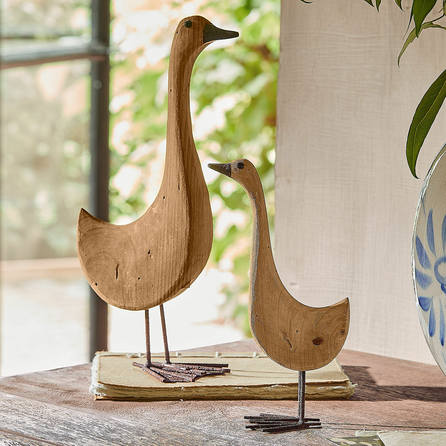 Deko-Gans 2er Set Amiraz Holzdekorationen in Vogel-Form in Naturfarbe auf Metallfüßen auf einem Tisch platziert.