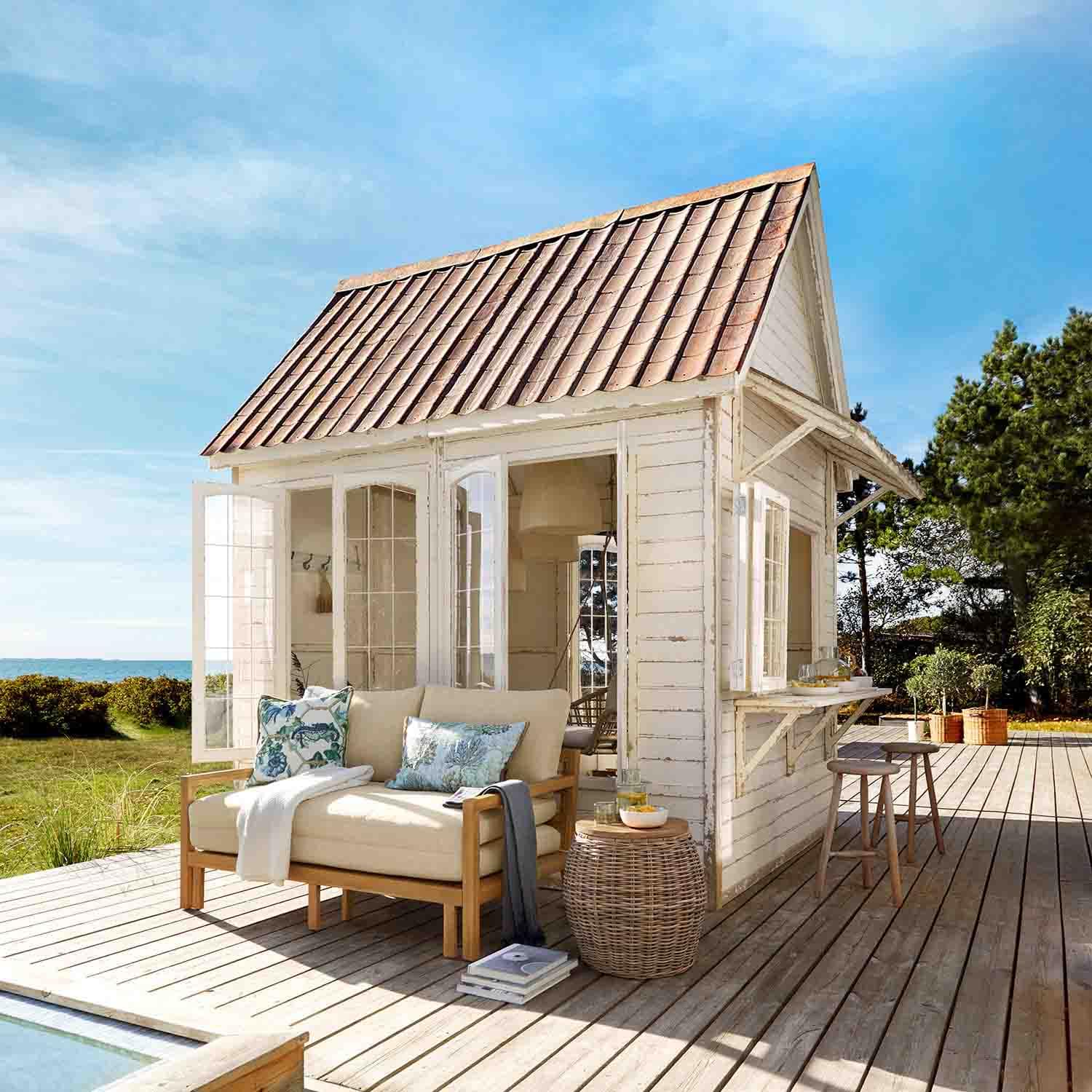 Weißes Holzhaus mit rotem Dach auf Holzterrasse mit beigem Sofa und Korbtisch