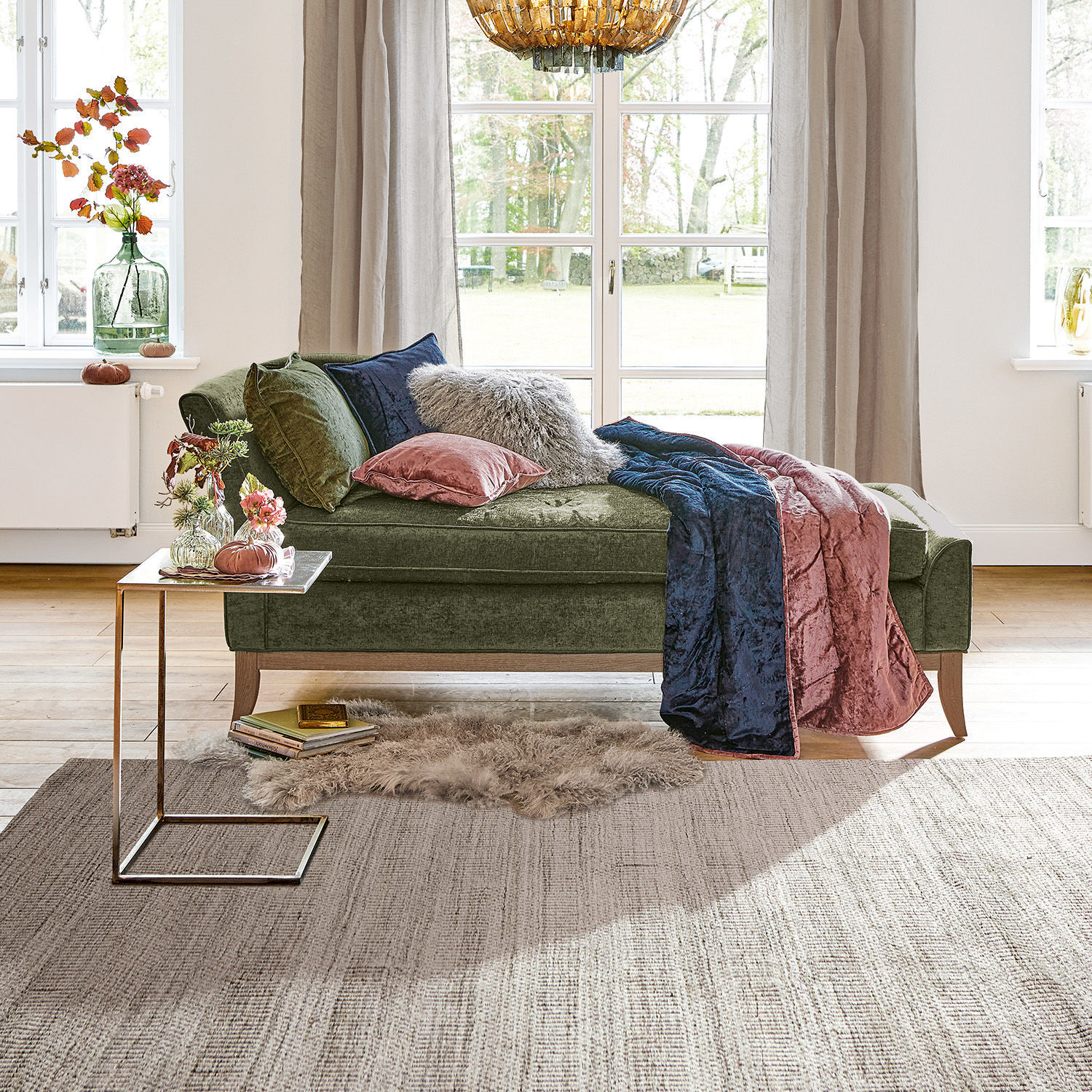 Chaiselongue Alphonse Grüne Samt-Chaiselongue mit Kissen und rosa-blauer Decke in hellem Wohnzimmer mit großem Fenster