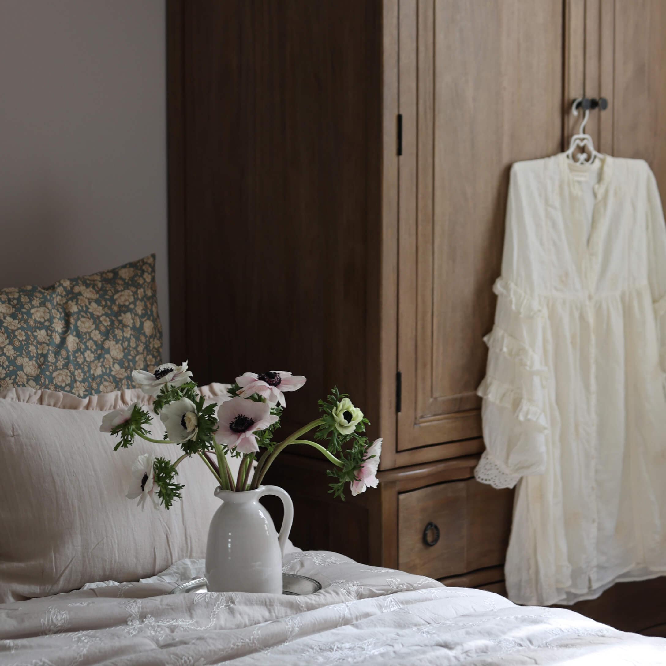 Weißer Keramikkrug mit Blumen auf hellem Bett vor einem Holzschrank im Schlafzimmer