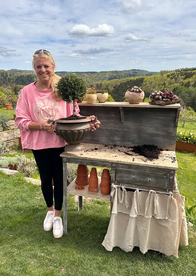 Frau in rosa Pullover hält Topf mit grünem Strauch an altem Gartentisch vor grünem Landschaftshintergrund