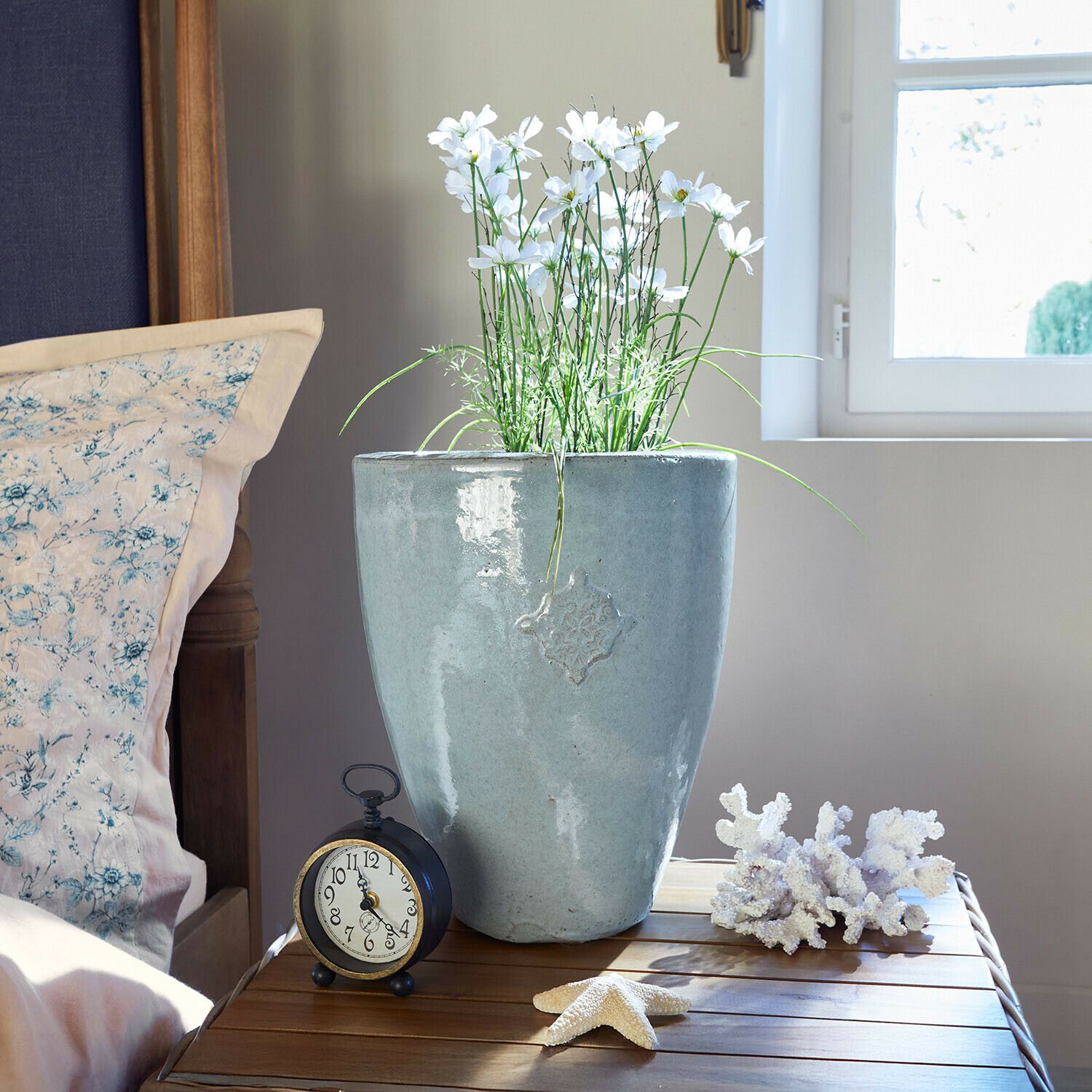 Übertopf Serenzia Grauer Keramikblumentopf mit weißen Blumen auf Holztisch neben Wecker und Meeresdekorationen