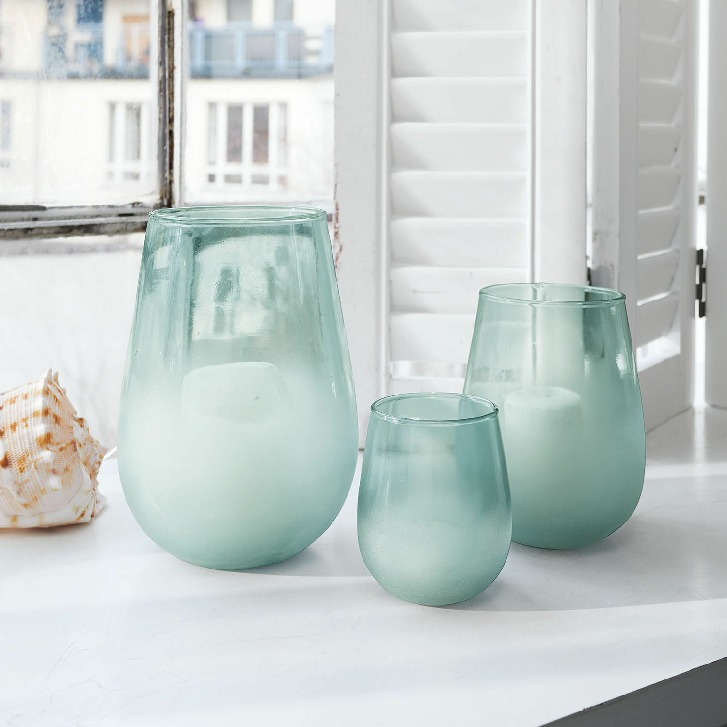Windlicht 3er Set Miristelle Drei matte grünliche Glaswindlichter auf weißem Fensterbrett mit Kerzen im Inneren