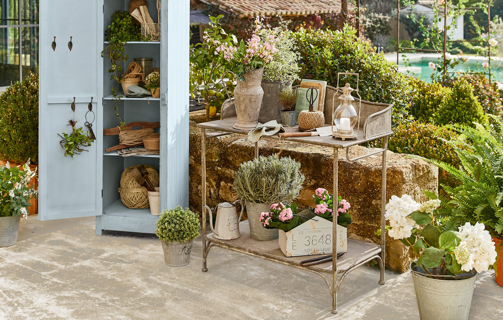 Metall-Gartenwagen mit Shabby-Chic-Dekorationen und Blumen auf Terrasse neben blauem Schrank.