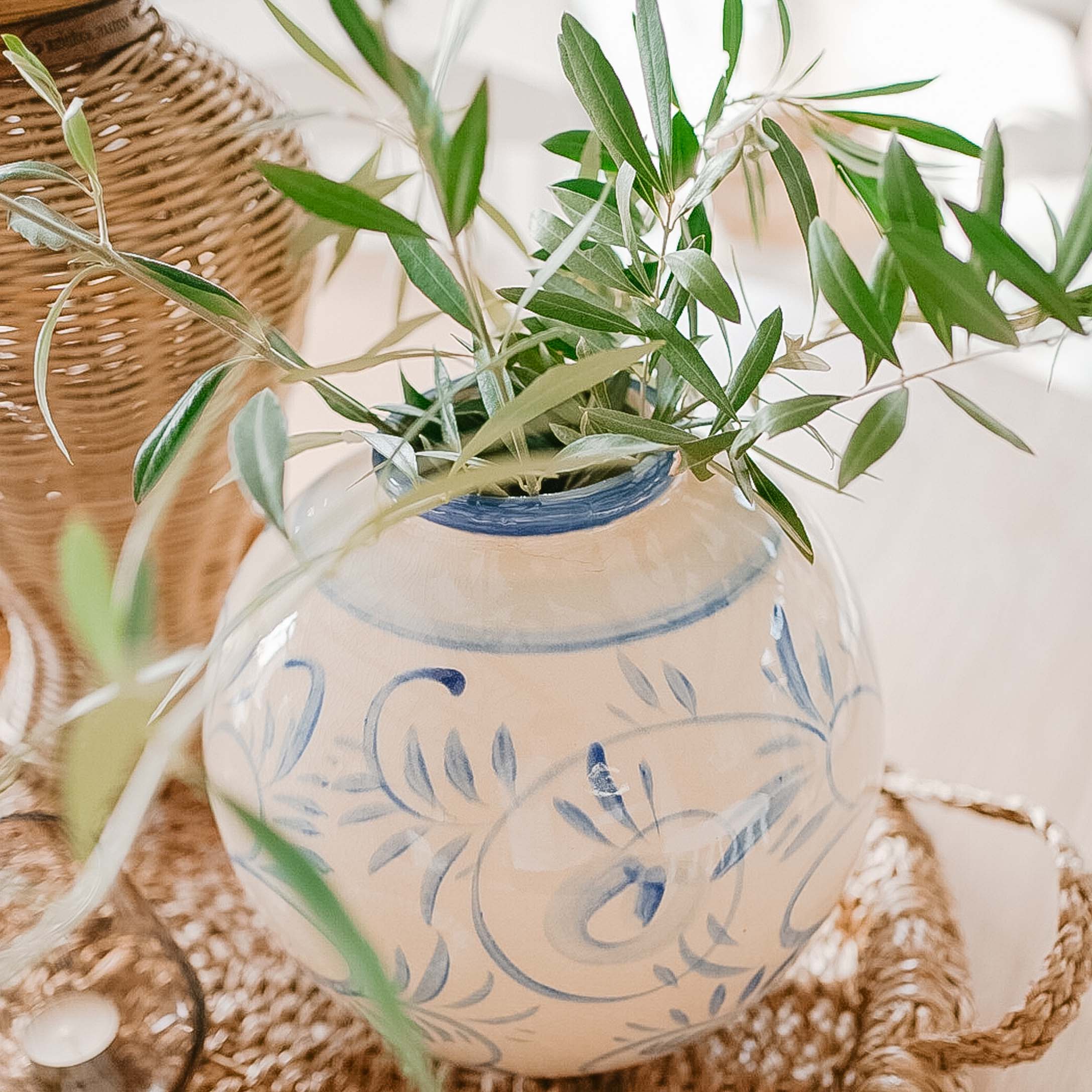 Weiße Keramikvase mit blauem Muster und grünen Blättern auf einem geflochtenen Tablett, Frühlingsdeko