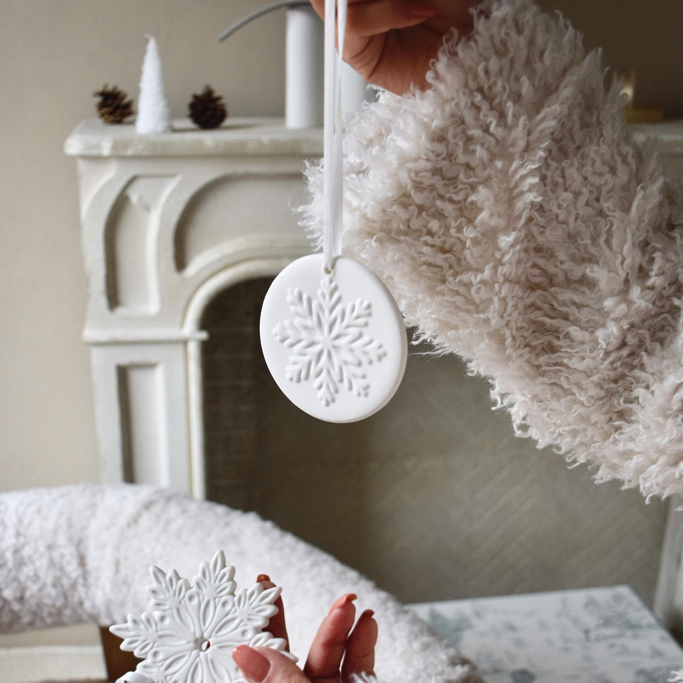 Weiße Keramik-Weihnachtsanhänger mit Schneeflockenmotiv vor Kamin und flauschigem Pullover