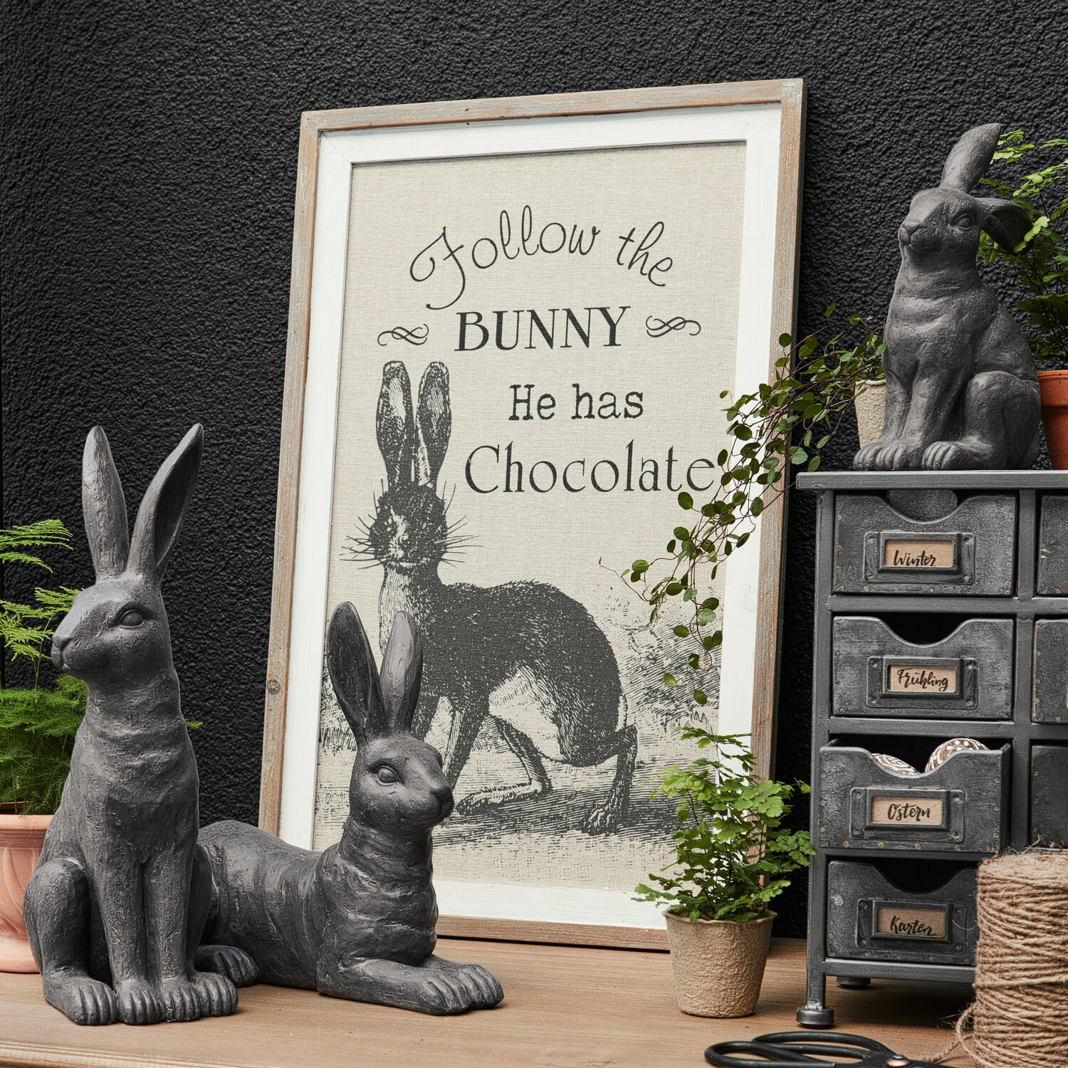 Dekoboard Chocolate Graue Hasenfiguren aus Deko mit Rahmen und Schriftzug über Hasen und Schokolade auf Holztisch