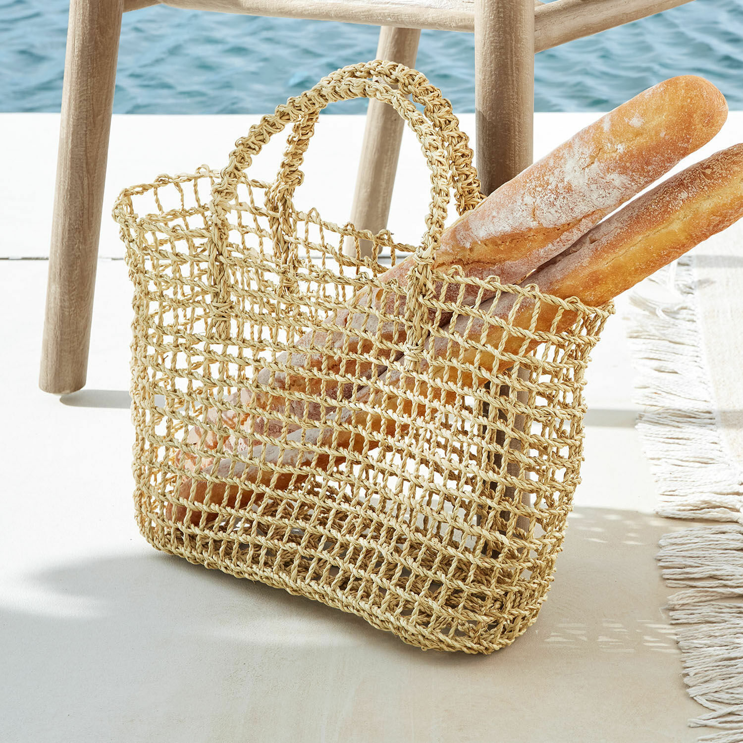 Tasche Xaintieux Natürliche Einkaufstasche aus geflochtenem Material mit zwei Baguettes neben einem Holzstuhl