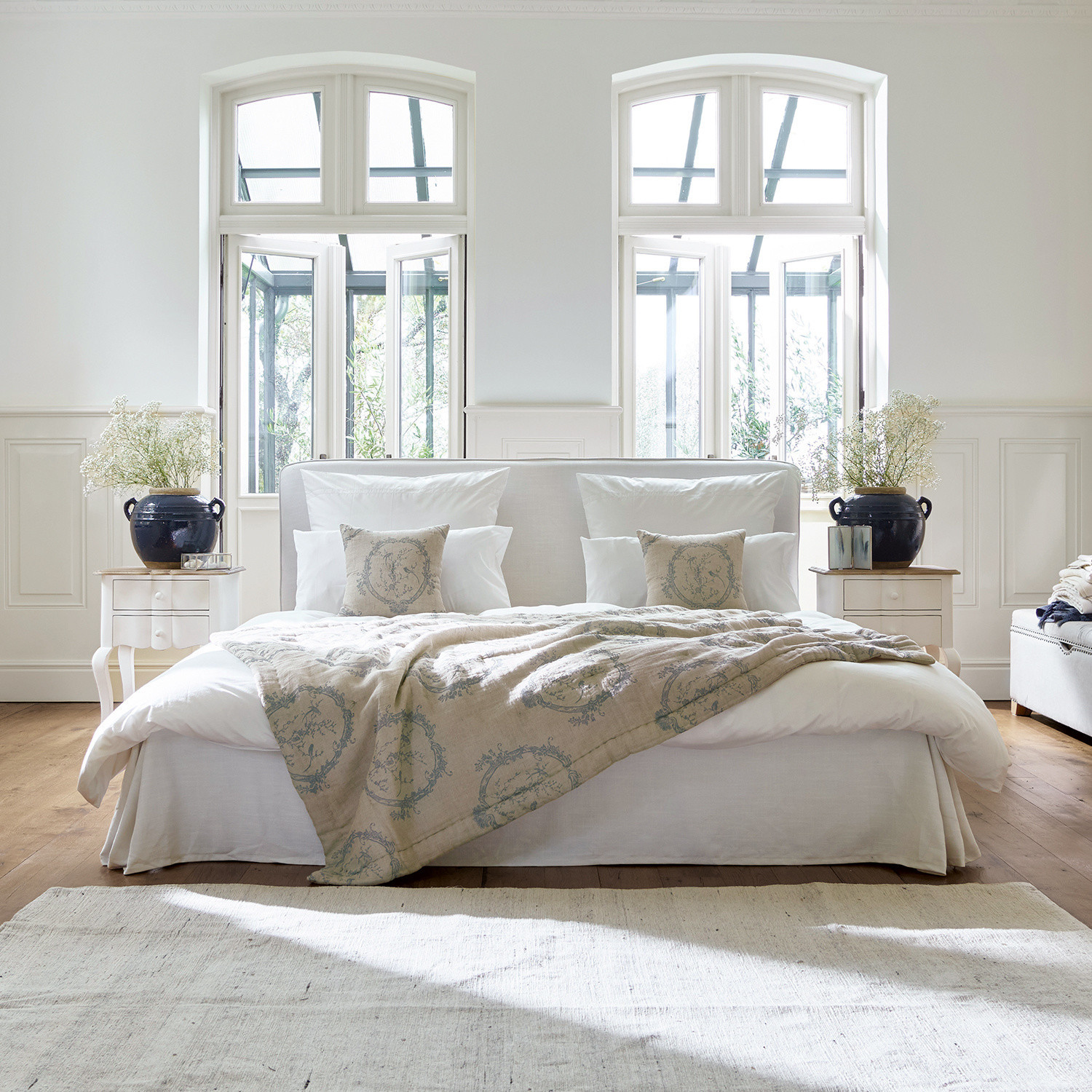 Bett Aubrianne Gemütliches Bett mit hellem Bezug und beiger Decke mit Muster in hellem Schlafzimmer mit großen Fenstern