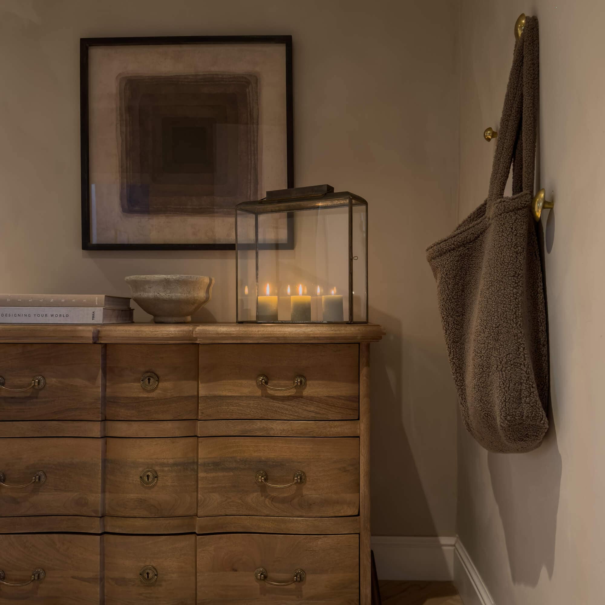 Holzkommode mit Messinggriffen, Glaslaterne mit brennenden Kerzen und braune Tasche aus weichem Material an der Wand
