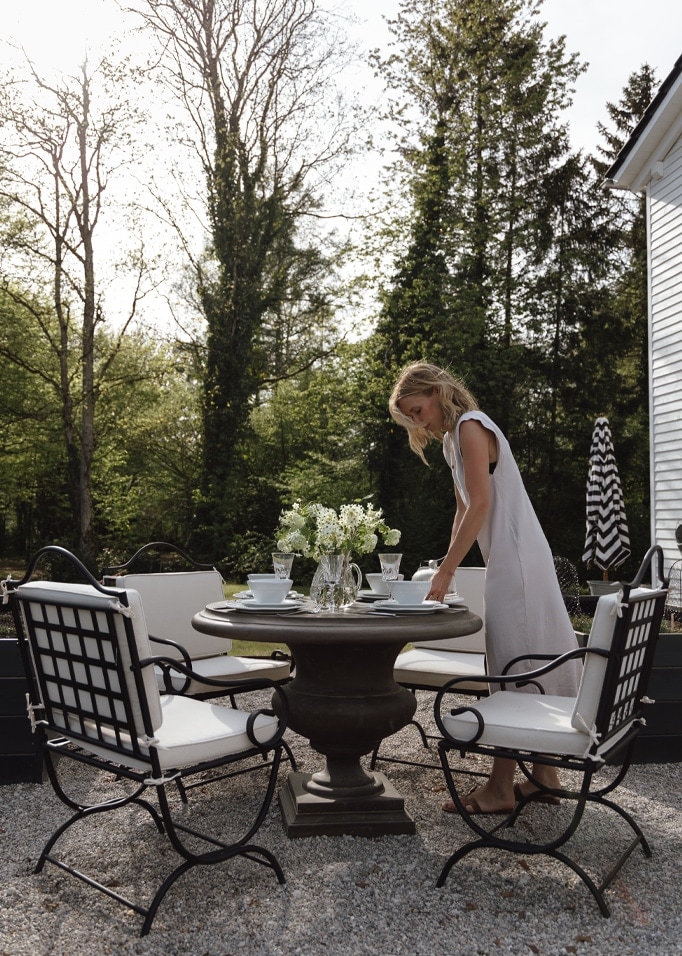 Garten mit rundem Steintisch und schwarzen Metallstühlen mit weißen Polstern, Frau im weißen Kleid.