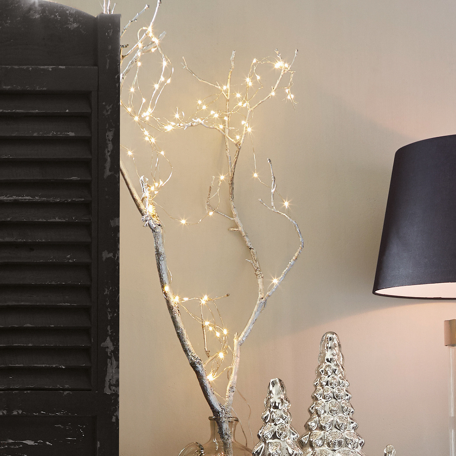 Lichterkette Loperhet Dekorativer Ast mit LED-Lichterkette in klarem Glasvase neben silbernen Weihnachtsdeko und schwarzer Lampe