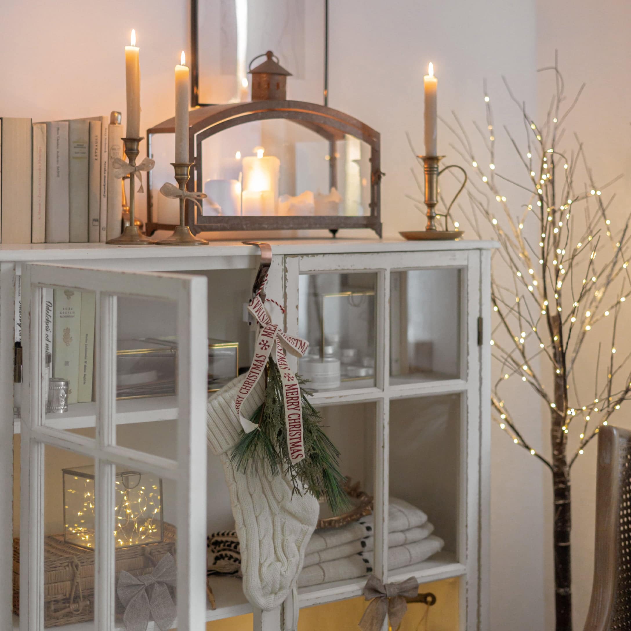 Weiße gestrickte Weihnachtssocke mit grünen Zweigen an weißem Schrank mit Kerzen und Lichterzweig