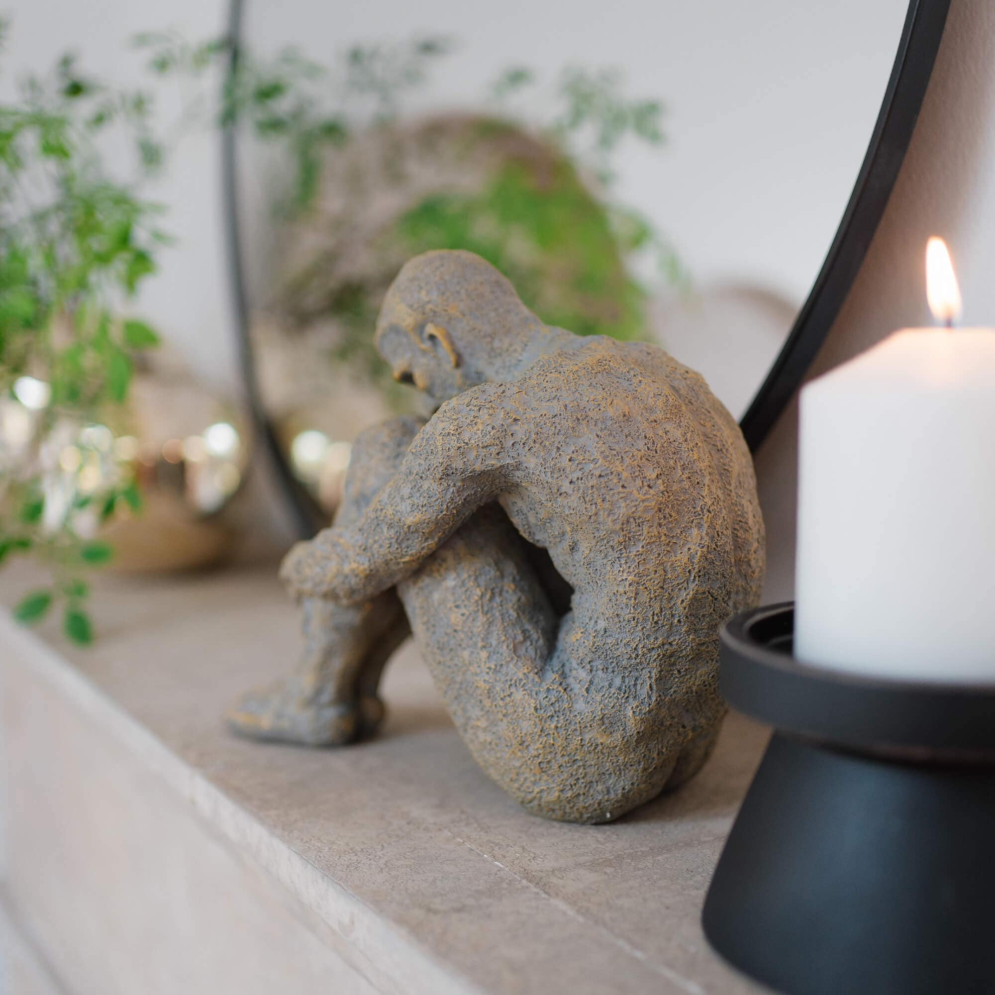 Graue Steinskulptur einer sitzenden Figur auf Marmorablage neben weißer Kerze