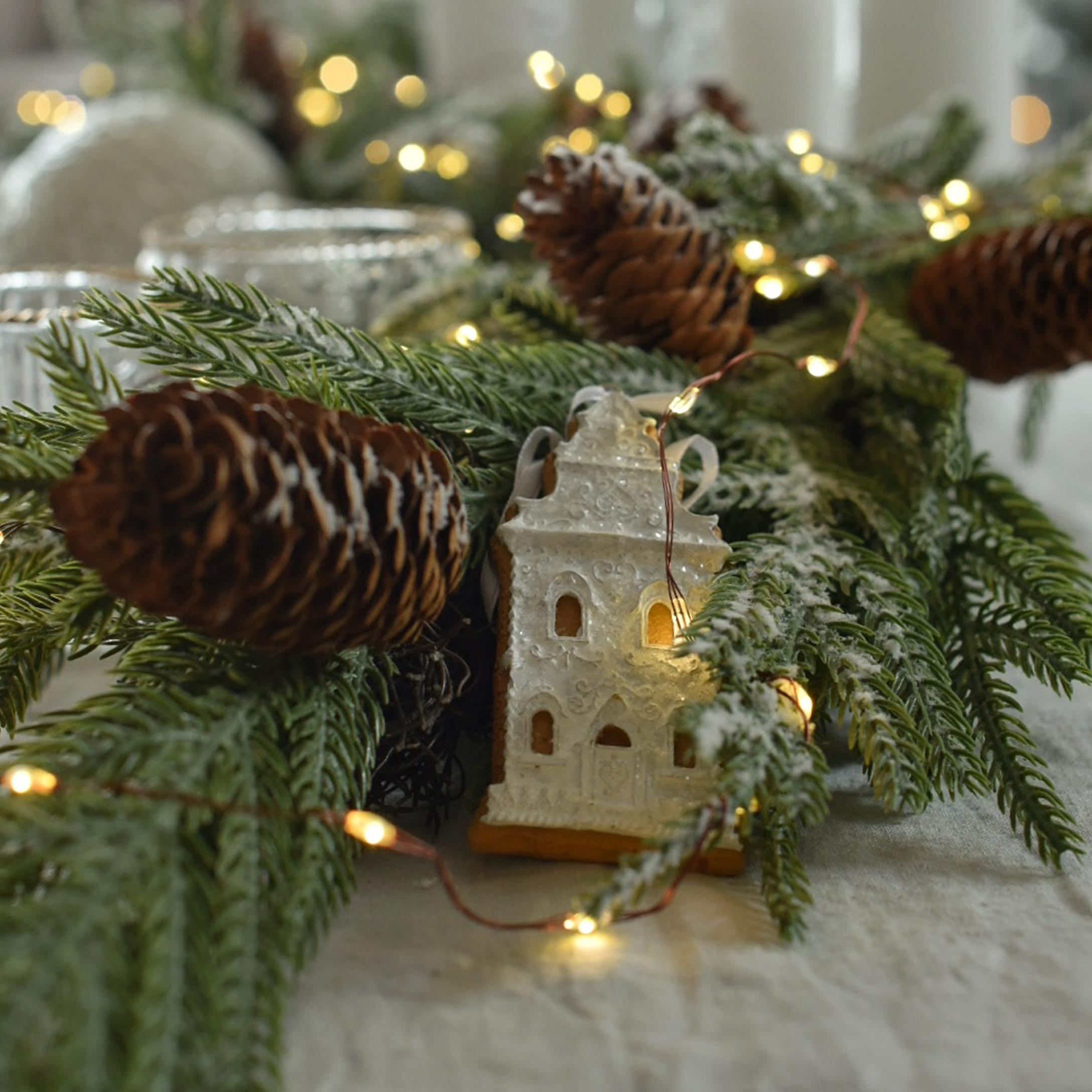 Weihnachtsdekoration mit grünen Zweigen, Tannenzapfen und beleuchtetem Lebkuchenhaus auf Tisch