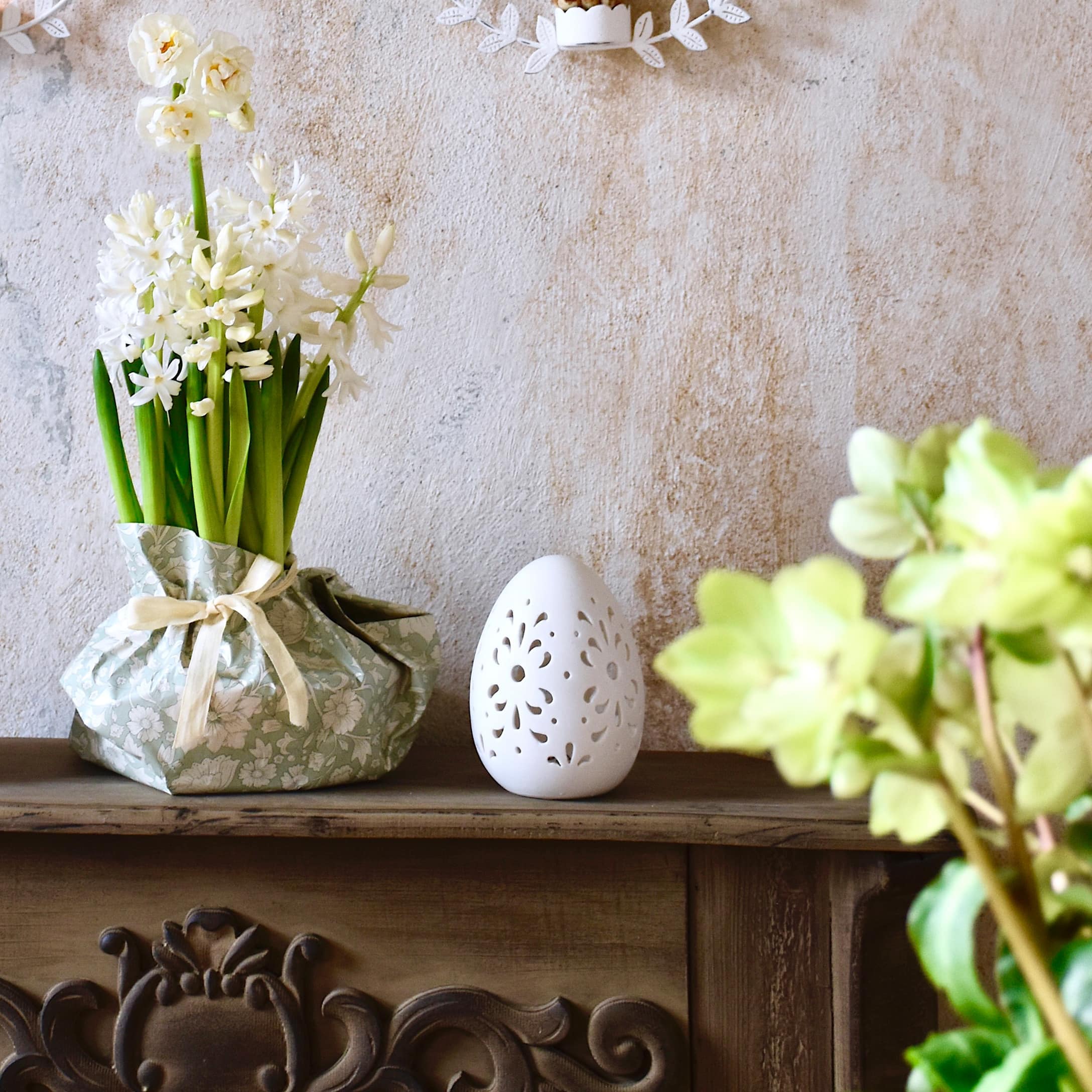 Weiße Blumen in grünem dekorativem Papier und weiße Keramik-Eierdekoration auf Holzregal