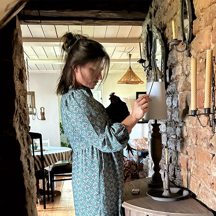 Frau im blau gemusterten Kleid zündet Lampe in rustikalem Raum mit Backsteinwand und Kerzen an