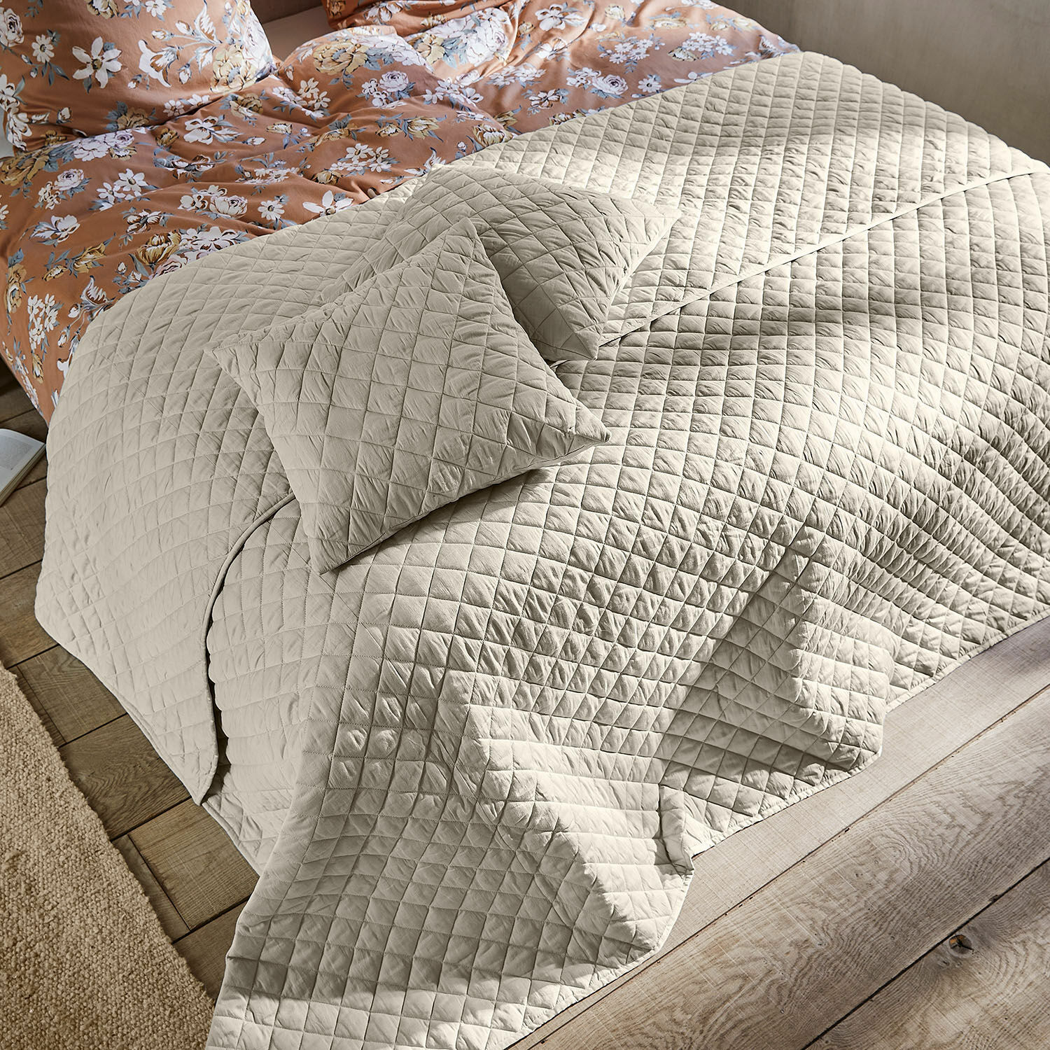 Quilt Jolique Beige gesteppte Tagesdecke und Kissen auf Bett mit Holzboden und geblümter Bettwäsche